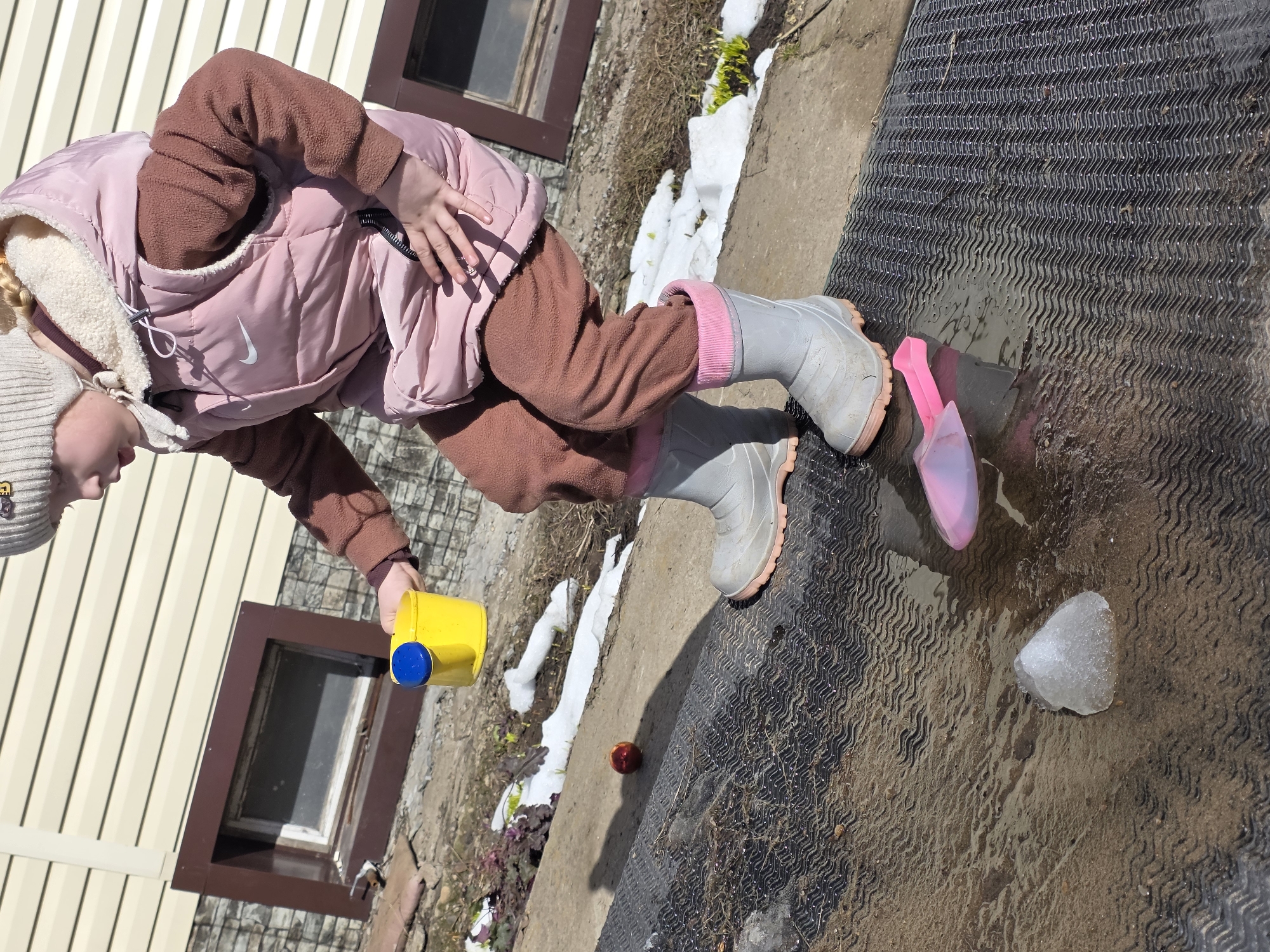 In Chaikovsky, Russia, a young child is depicted outdoors during what appears to be late winter or early spring. The child, wearing a light beige knit beanie, a pink puffer vest with a white Nike logo, a brown fleece top, brown fleece pants, and light grey rubber boots with pink accents, is crouching low. Their right hand holds a yellow plastic watering can with a blue top. A single piece of clear ice, roughly triangular, rests on a wet, dark grey corrugated rubber mat in the foreground. Nearby on the mat, a pink plastic scoop lies flat. Behind the child's left leg, a red apple is visible on a concrete edge bordering a dirt area containing remnants of snow and some dry vegetation. The background features the exterior wall of a building, comprising light beige horizontal siding above a dark brown section with what appears to be a stone or brick foundation. Two grimy, rectangular basement windows with dark brown frames are visible in the lower part of the wall. The scene is brightly lit by sunlight, casting distinct shadows.
