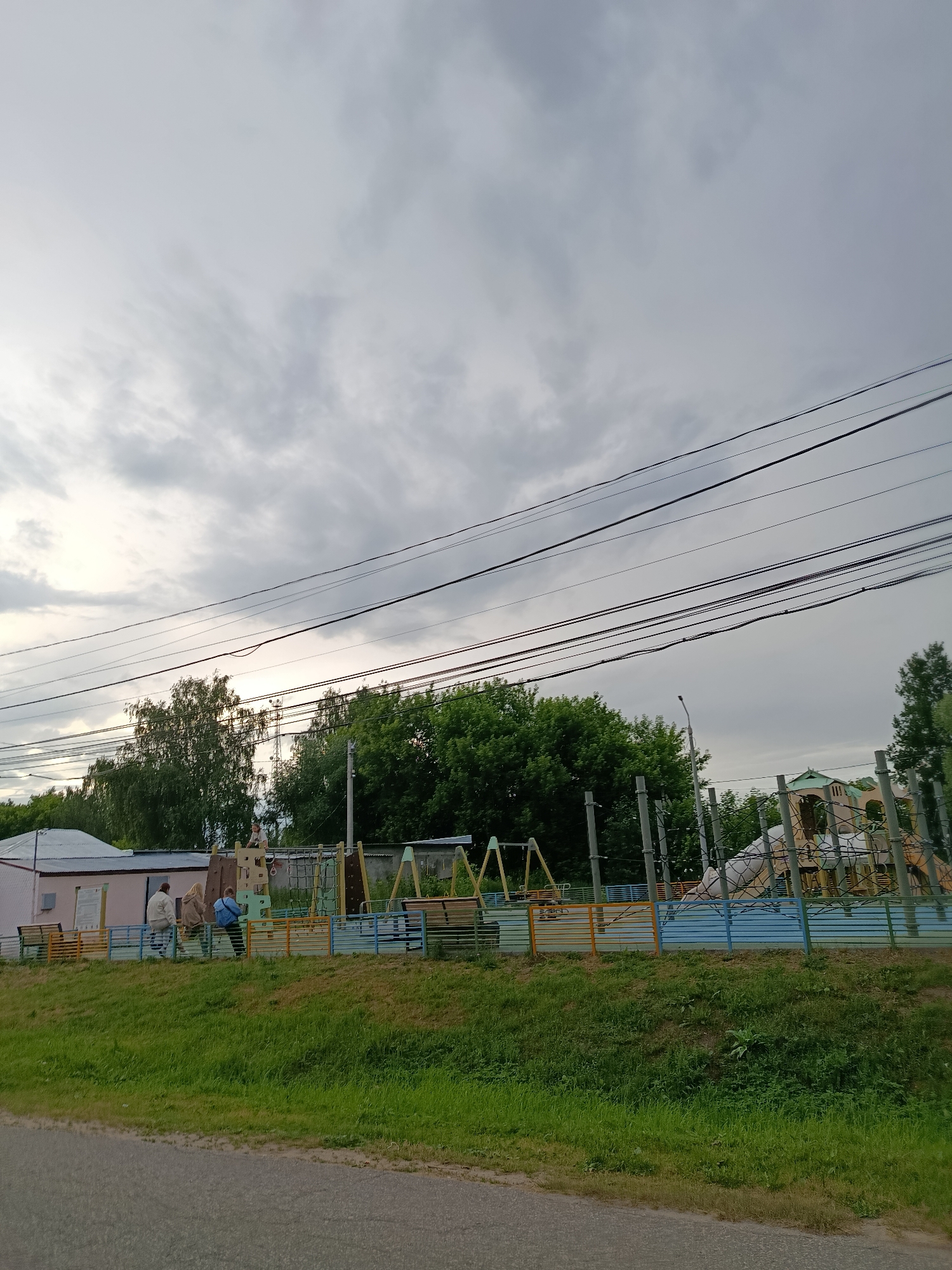 The image depicts a cloudy day scene in the city of Vereya, Russia, featuring a playground and some surrounding structures. In the foreground, a paved road runs horizontally, with a grassy embankment sloping upwards towards the playground. The playground itself is a colorful structure with blue and yellow fencing. It includes swings and a climbing net. To the left of the playground, a low, pink building with a white roof is visible, with benches in front of it. Three individuals are present: two standing near the building and one sitting on the playground structure. One person appears to be wearing a blue jacket, another a light brown coat, and a third person is partially obscured. In the background, dense green trees form a backdrop, and overhead, multiple power lines are strung across the sky. The overall atmosphere is overcast and somewhat subdued, typical of a cloudy day.