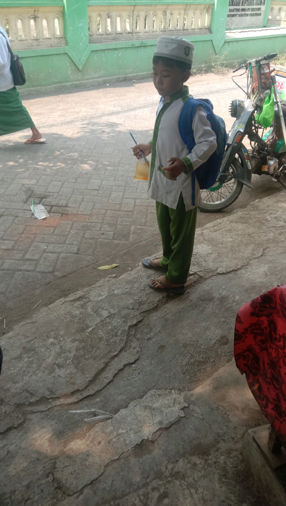 In Ngempit, Indonesia, a young boy wearing a white skullcap, white shirt with green trim, and green pants stands on a paved walkway. He carries a blue backpack and holds a plastic bag containing a yellow beverage with a straw. To his right, a motorcycle is parked. In the background, a green building with white decorative elements is visible. Another person, partially visible on the left, wears a white shirt and a long green skirt, with a black bag slung over their shoulder. The ground is a combination of paved brick and rough concrete.