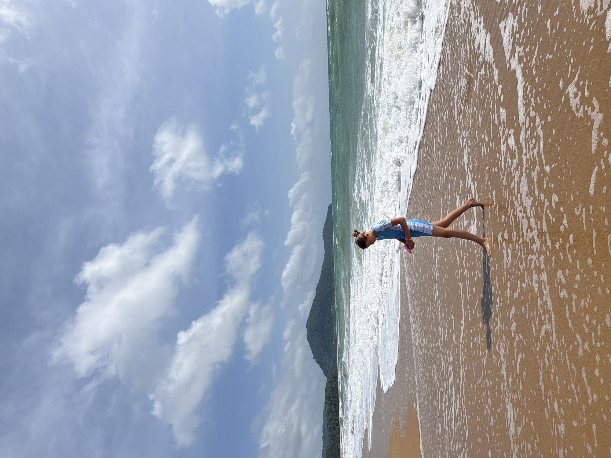 In Mai Khao, Thailand, a young person wearing a blue and white patterned swimsuit walks along a sandy beach. The water is a deep green, with white waves crashing onto the shore. The sky is a bright blue, dotted with fluffy white clouds. In the distance, a lush green mountain range can be seen. The sand is wet and reflects the sunlight, with small waves of foam washing over it. The person is holding a pink object in their right hand.