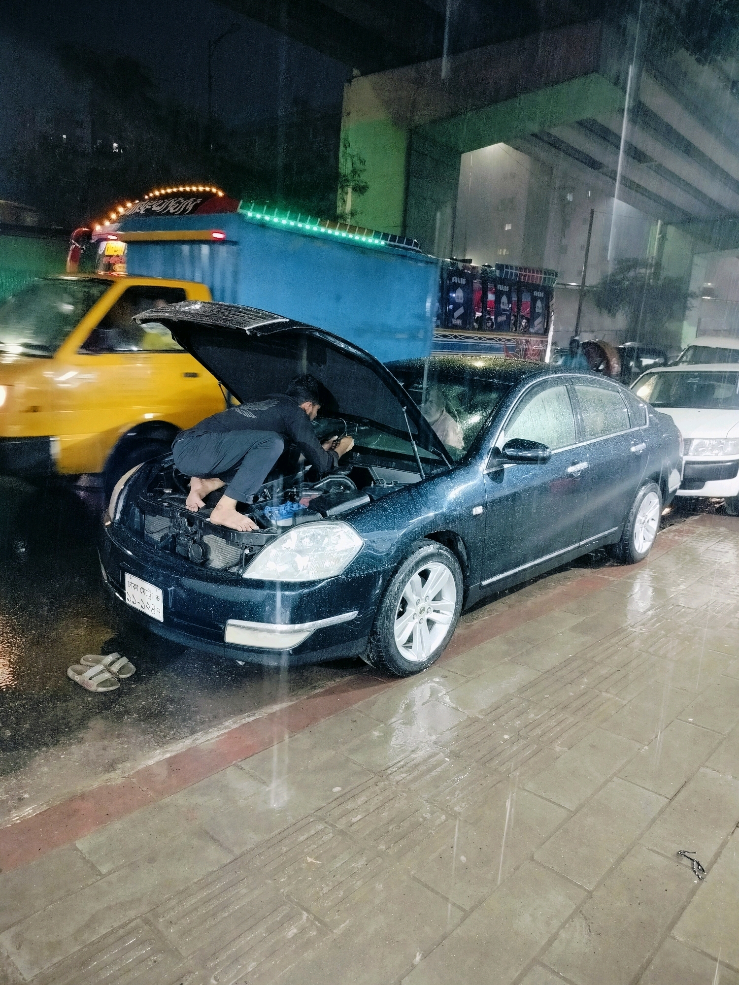 A dark sedan is parked on a wet street in Dhaka, Bangladesh, with its hood open. A person, dressed in dark clothing and barefoot, is crouched over the engine, appearing to be working on it. The street is wet with rain, and raindrops are visible falling. A yellow vehicle is partially visible in the background to the left. Other vehicles and a building are also visible in the background. A pair of sandals lies on the wet pavement near the front of the dark sedan. The scene appears to be at night or under heavy cloud cover, with artificial lights illuminating the area.
