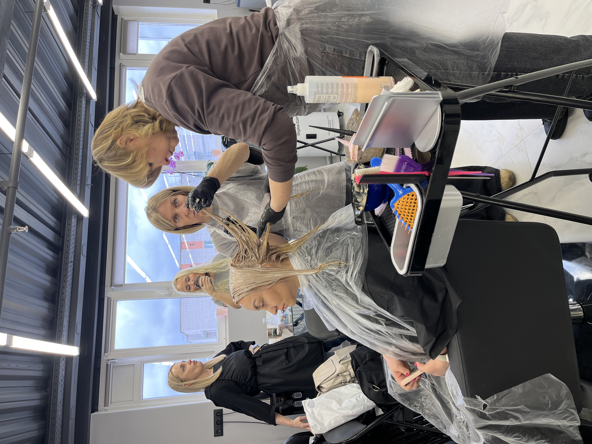 The image captures an active scene inside a modern hair salon in Izhevsk, Russia, during what appears to be daytime, given the natural light from the windows and cloudy sky.
The central activity involves two women, presumably hairdressers, meticulously applying product to the blonde hair of a client who is seated, wearing a clear plastic cape. One hairdresser, wearing a brown top and black gloves, leans over the client, while another, also in a light top and black gloves, assists. The client receiving treatment has a clear plastic band around her head.
A rolling salon trolley, laden with various hair products, brushes (some branded "K-COLOR"), combs, clips, and bowls, is positioned next to the client. A prominent white bottle with an orange label, labeled "Digital Color," sits on the trolley.
In the background, another blonde woman, wearing a light grey top, stands holding a cup and smiling, observing the process. In the foreground, towards the bottom left, a fourth blonde woman, dressed in a black top or apron, is seated in a salon chair, with a beige handbag and a white plastic bag nearby.
The salon features a contemporary design with black corrugated walls or ceiling panels, fluorescent tube lighting, and white window frames. A potted orchid can be seen on a windowsill, adding a touch of nature to the interior. The overall atmosphere suggests a professional and busy salon environment.