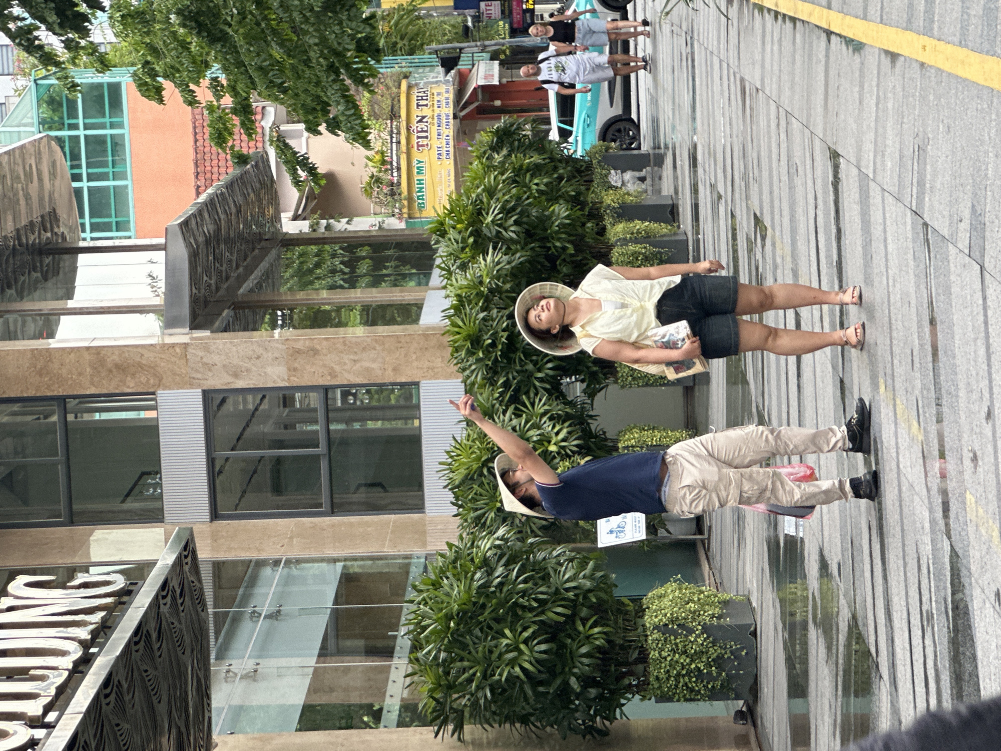 In Đà Nẵng, Vietnam, a man in a blue t-shirt and beige cargo pants, wearing a conical hat, points upwards with his right hand. He stands near large potted green plants on a wet, paved sidewalk. Beside him, a woman in a cream-colored, open-necked shirt, denim shorts, and a conical hat looks upwards, holding a bag with a magazine. In the background, a turquoise car is parked, and two people, a man and a woman, walk away from the camera. Buildings with glass windows and balconies line the street, with signs for "BÁNH MỲ TIÊN THÁI" visible. The sidewalk is wet, reflecting the overcast sky.