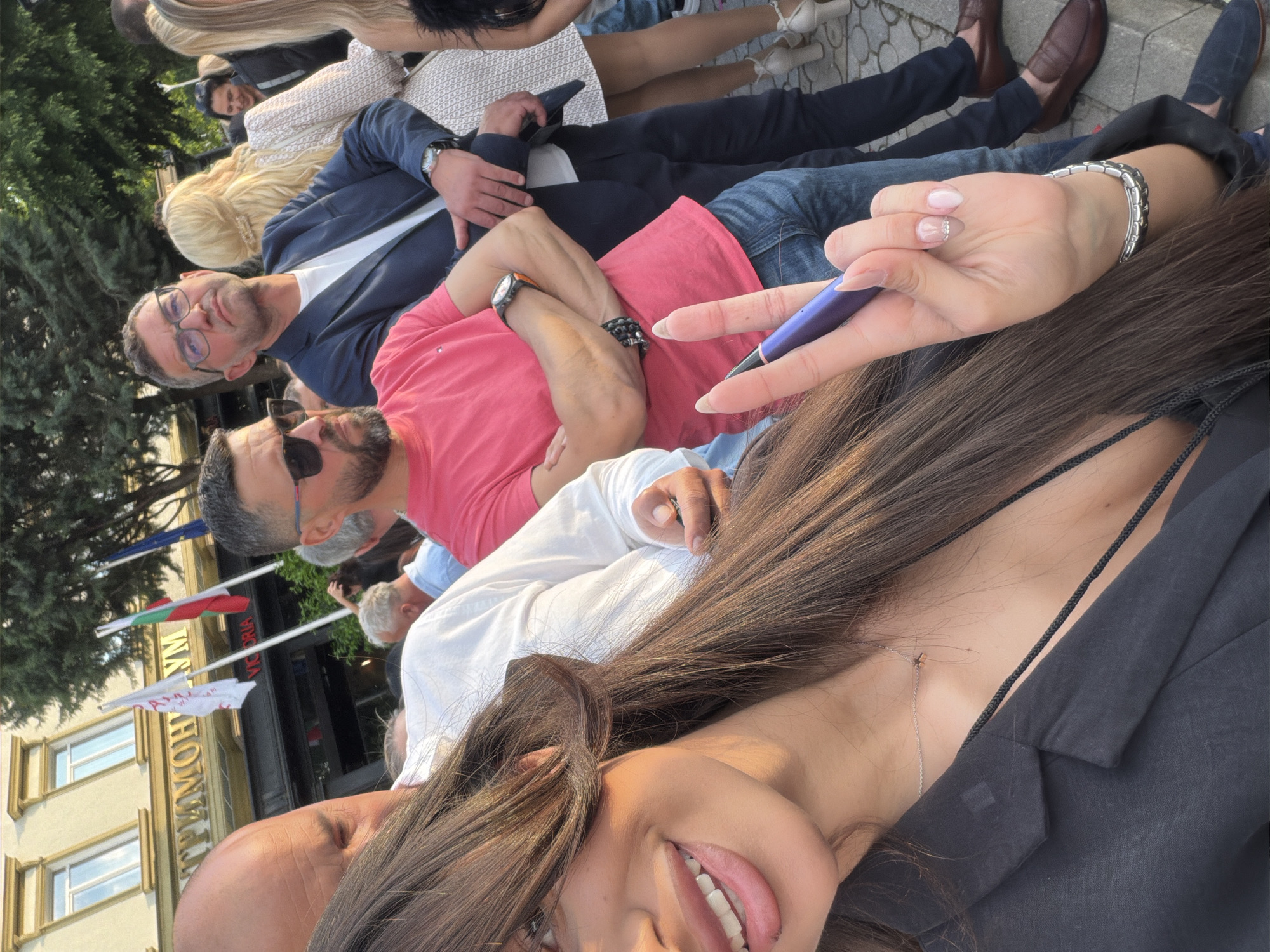 A selfie taken in Plovdiv, Bulgaria, features a young woman with long brown hair smiling at the camera with her tongue slightly out. She is wearing a black blazer and a delicate silver necklace. Her manicured hand holds a blue pen or stylus.
Behind her, several people are visible. One man wears glasses and a navy blazer. Another man wears sunglasses and a salmon-colored shirt. Other individuals are partially visible.
In the background, flags of Bulgaria and the European Union are displayed. A building with "GRAND HOTEL TRIMONTIUM" written on it is also present, suggesting the photograph was taken near this landmark in Plovdiv. The scene appears to be outdoors during daylight.