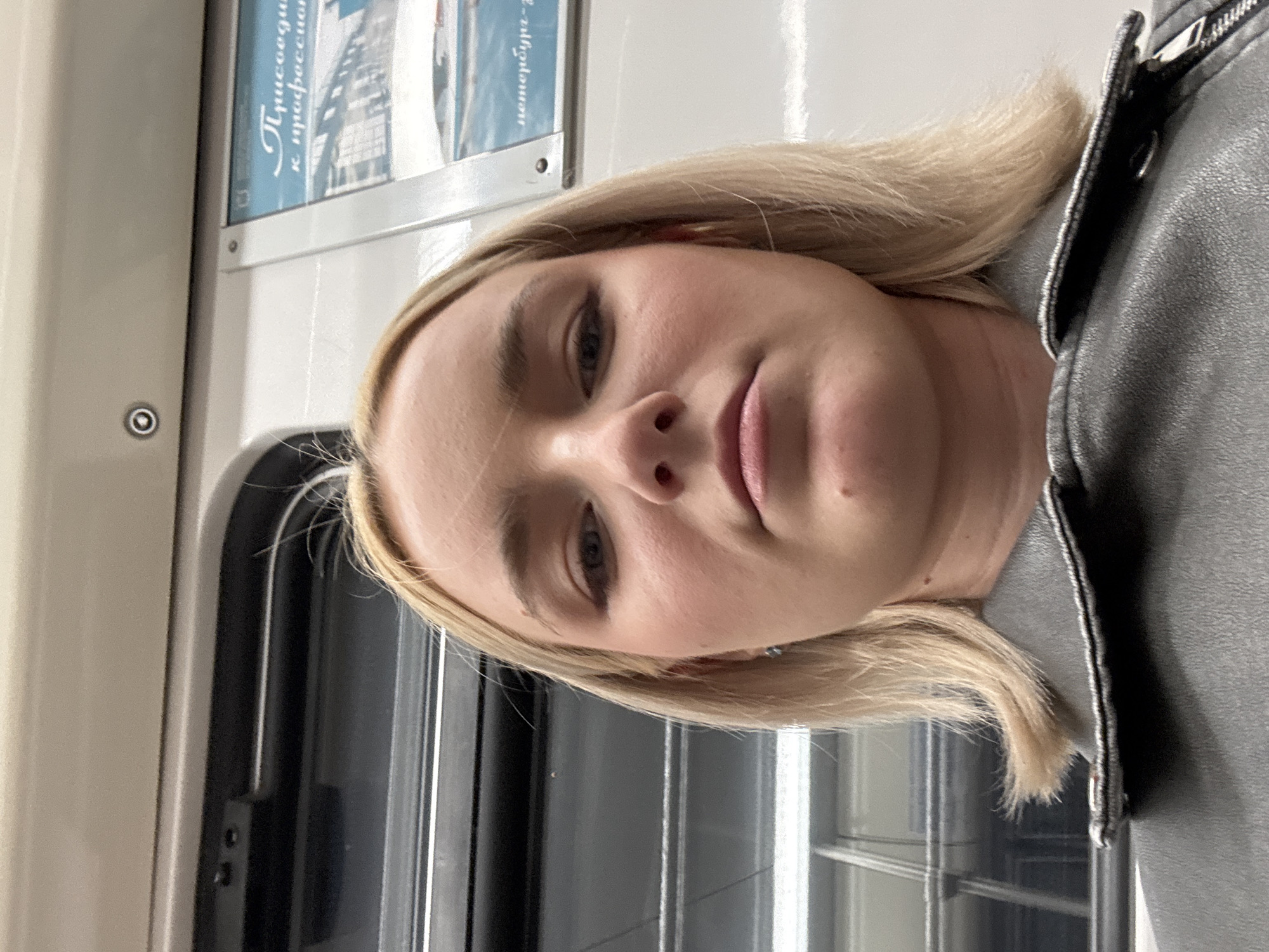The image is a selfie taken by a young Caucasian woman with blonde hair, wearing a gray leather jacket. She is looking directly at the camera with a neutral expression. Her makeup includes eyeliner and mascara. The background appears to be the interior of a subway car, with a window and a poster visible. The lighting is somewhat dim, suggesting an indoor environment. The location is unknown, and the time of day is also not specified.