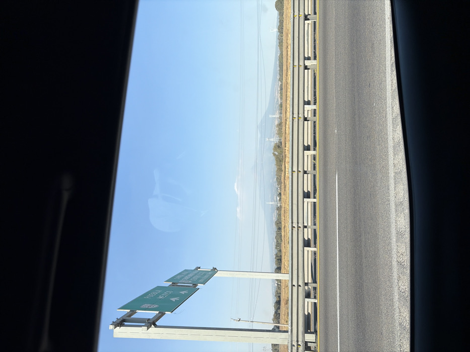 A view from a car window shows a highway under a clear blue sky. Guardrails run along the side of the road. A green highway sign indicates directions to Puebla and Mexico, both marked with arrows. Distant mountains are visible on the horizon, with power lines stretching across the landscape.