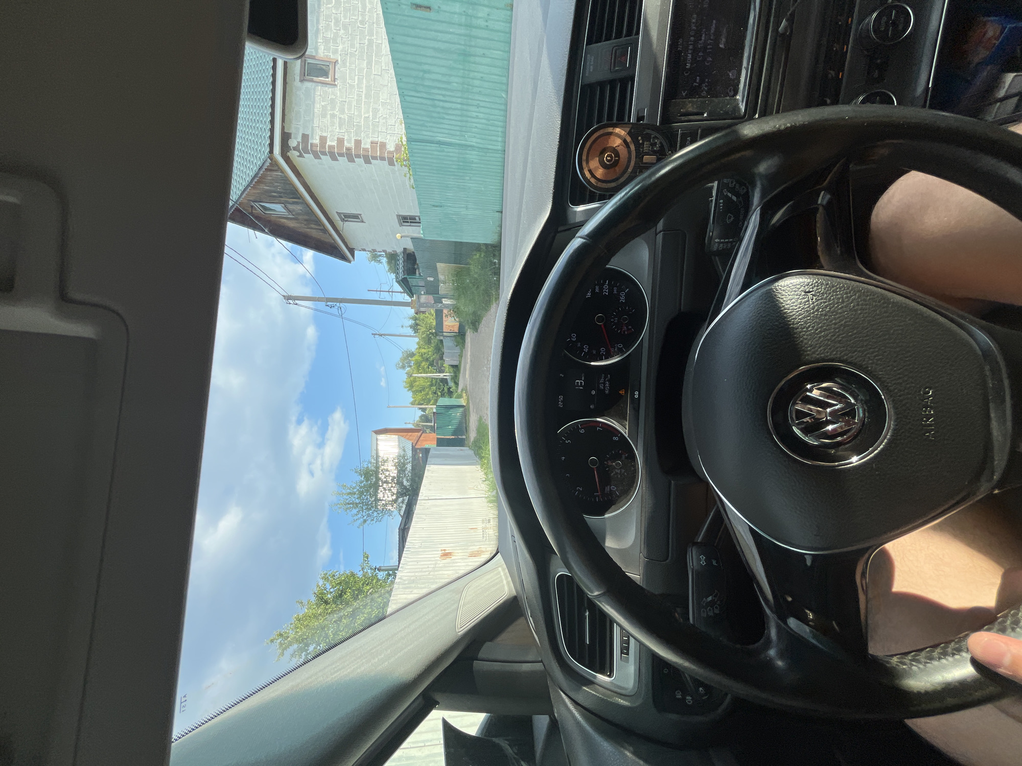 A first-person view from inside a car shows a residential street in Vasilyevo, Russia. The driver's hands are visible on the steering wheel, and the car's dashboard displays speed and other information. The street is lined with houses and fences, and the sky is blue with scattered clouds. The overall impression is of a quiet, everyday scene in a Russian town.