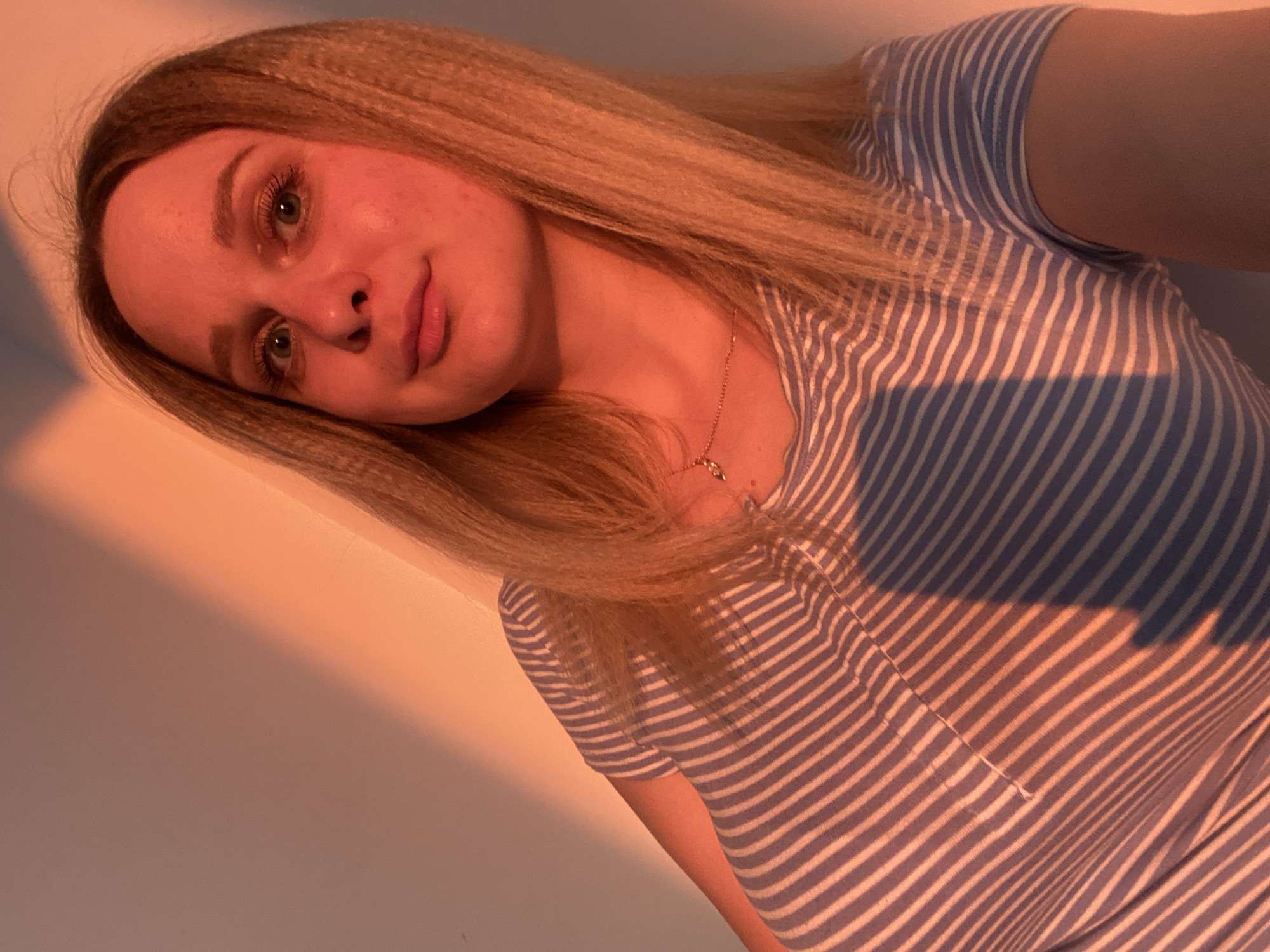 A young woman with blonde, crimped hair is photographed from the chest up in a selfie. She is wearing a blue and white striped t-shirt and a delicate necklace with a small pendant. The lighting suggests it is either sunrise or sunset, with warm orange light casting diagonal shadows across the wall behind her and onto her clothing. Her makeup is subtle, with mascara visible on her lashes. The background is a plain wall. There are no discernible landmarks to suggest a specific location within Izhevsk, Russia.