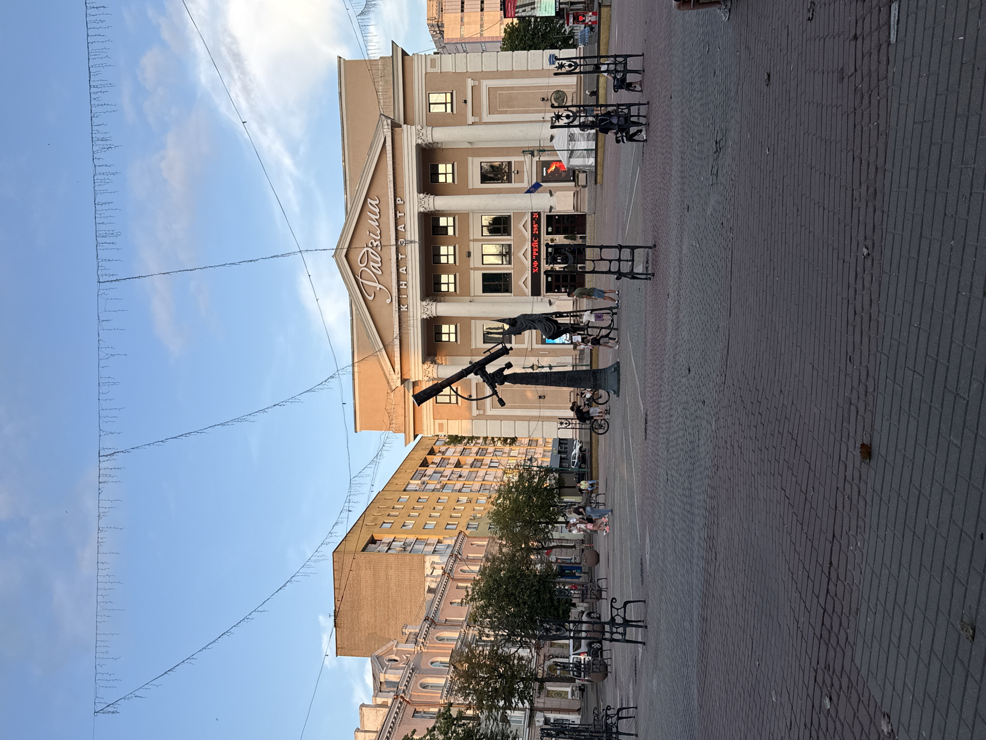 The image depicts a wide-angle view of a public square in Mahilyow, Belarus, likely in front of the "Radzima" cinema. The architecture suggests a mix of historical and Soviet-era styles. In the foreground, a cobblestone plaza is sparsely populated with people. Several unique, ornate metal chairs are scattered across the plaza, some occupied. A prominent sculptural element, resembling a telescope and a figure, stands near the center.
The "Radzima" cinema, a peach-colored building with classical columns and a pediment, dominates the background. Signage above the entrance indicates its name. To the left of the cinema, there are multi-story residential buildings with a mix of architectural details. Trees are present, suggesting a park-like or tree-lined urban setting. The sky is a clear blue with a few wispy clouds, and decorative string lights are suspended across the upper portion of the scene, indicating a festive or decorative lighting setup. The overall atmosphere appears to be during daylight, possibly late afternoon, given the lighting and soft shadows. People are engaged in casual activities such as walking, cycling, and sitting.