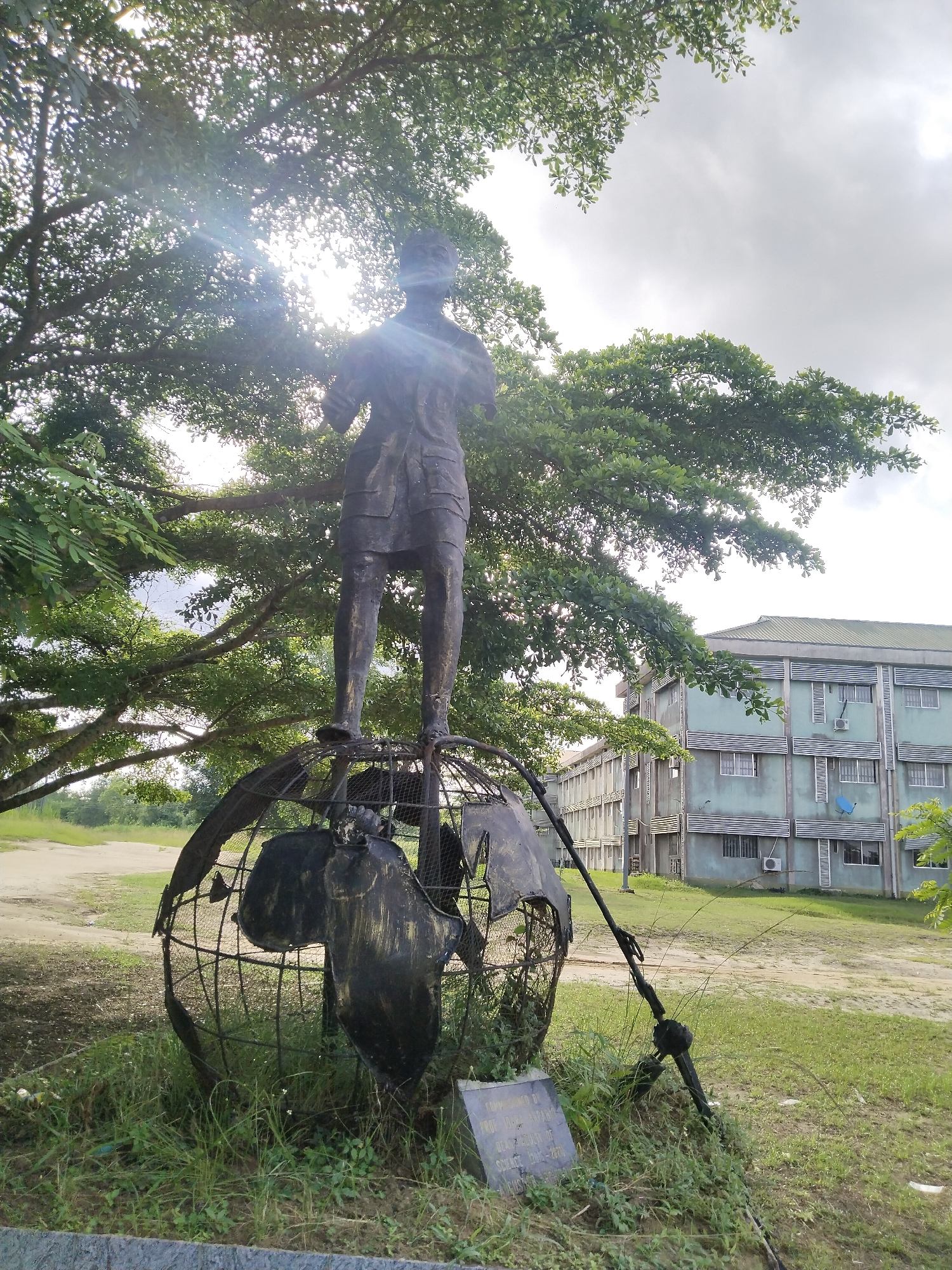 A statue of a person standing on a globe is centered in the frame. The globe is made of metal and the statue is dark. A plaque is at the base of the globe. Green trees are behind and around the statue. In the background is a multi-story building. The statue is located in the city of Uyo, Nigeria.