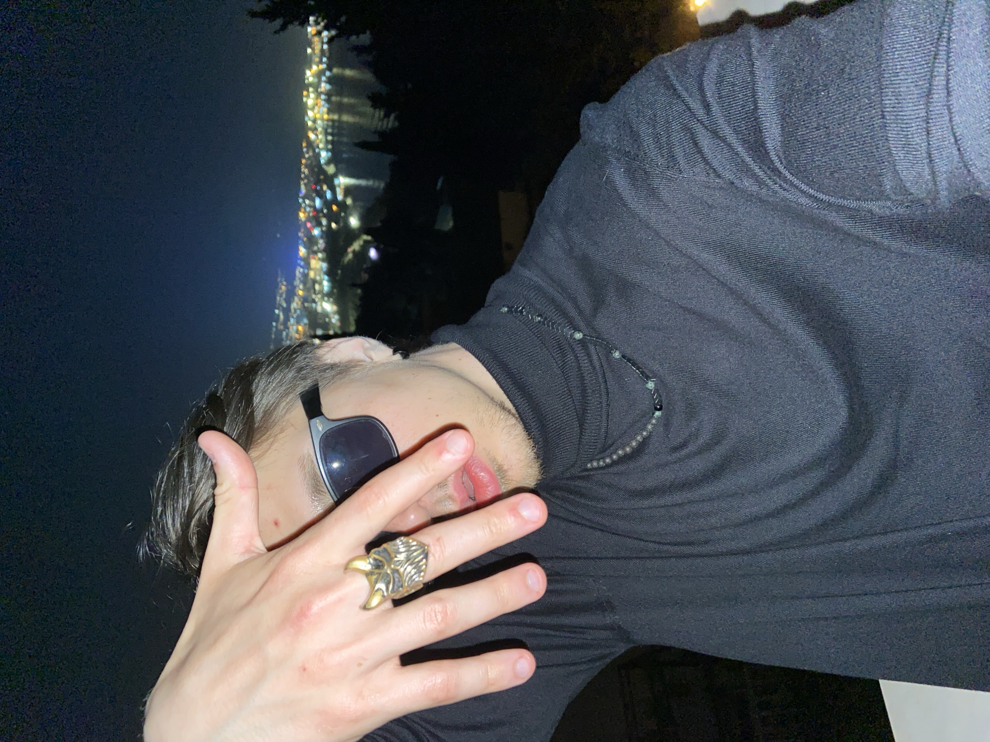 A person in Šušanj, Montenegro, wearing a black turtleneck shirt and sunglasses, stands outdoors at night with an illuminated cityscape in the background. The person's face is partially obscured by their left hand, which has a gold ring in the shape of an eagle's talons. A beaded necklace is visible around the person's neck.