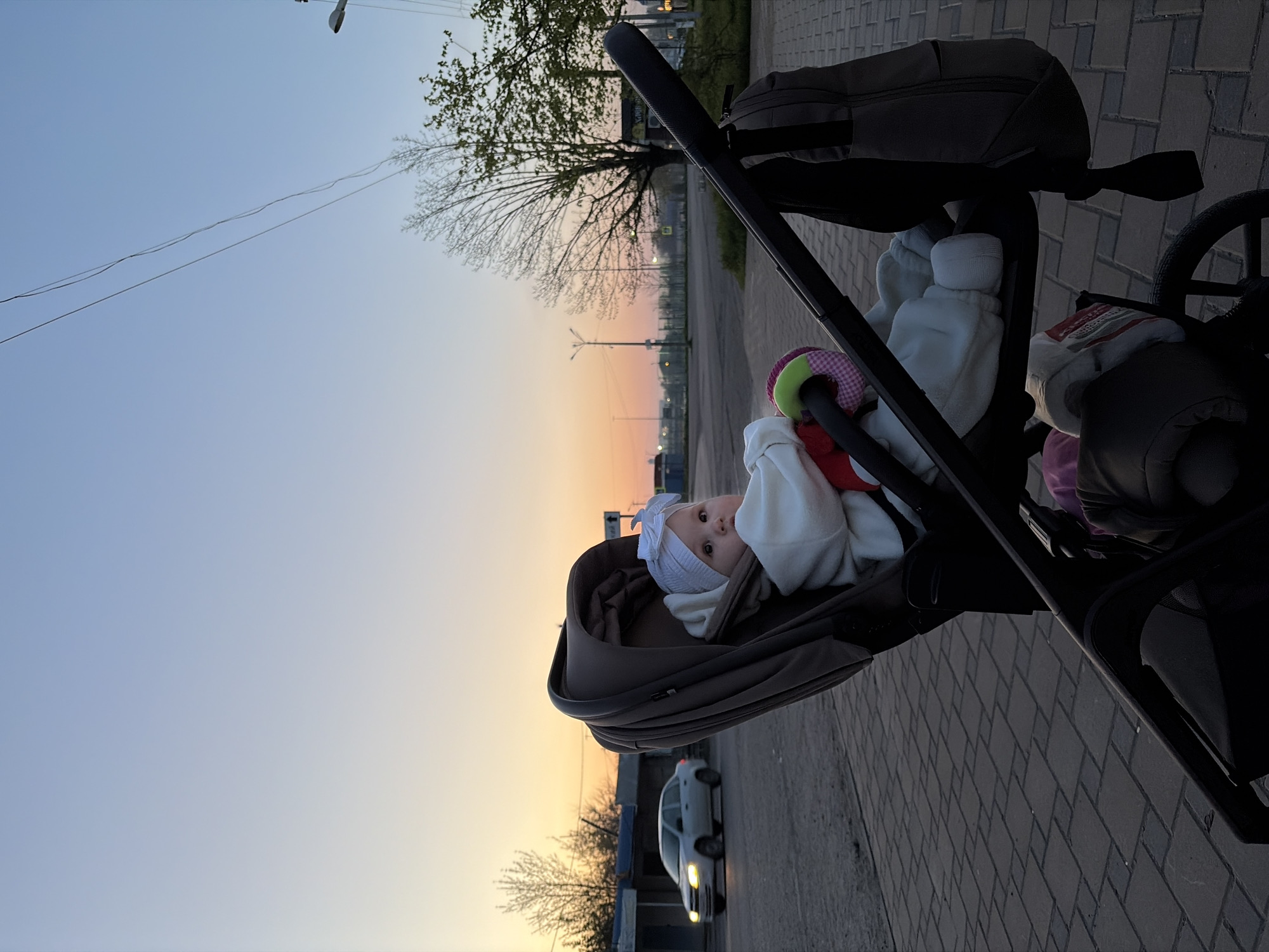 The image captures a baby in a stroller during sunset or dusk in Novopokrovskaya, Russia. The central subject is a baby, dressed in white clothing and a white hat with a bow, lying in a dark-colored stroller. The baby looks directly at the viewer with an alert and curious expression. A colorful soft toy is visible near the baby's hands.
The scene is outdoors, likely on a paved sidewalk or path alongside a road. The ground features a distinct block or brick pattern. In the background, trees with sparse foliage and some new green leaves are visible, suggesting springtime. Utility poles with wires stretch across the upper part of the frame. A white car with its headlights on is driving on the road in the mid-ground. Distant streetlights and vague building outlines are discernible along the horizon.
The time of day is clearly indicated by the sky, which transitions from a warm orange-yellow glow near the horizon to a soft blue higher up, characteristic of sunset or twilight. The weather appears clear with no visible clouds. The overall ambiance is calm and peaceful. No clearly legible text is visible in the image.