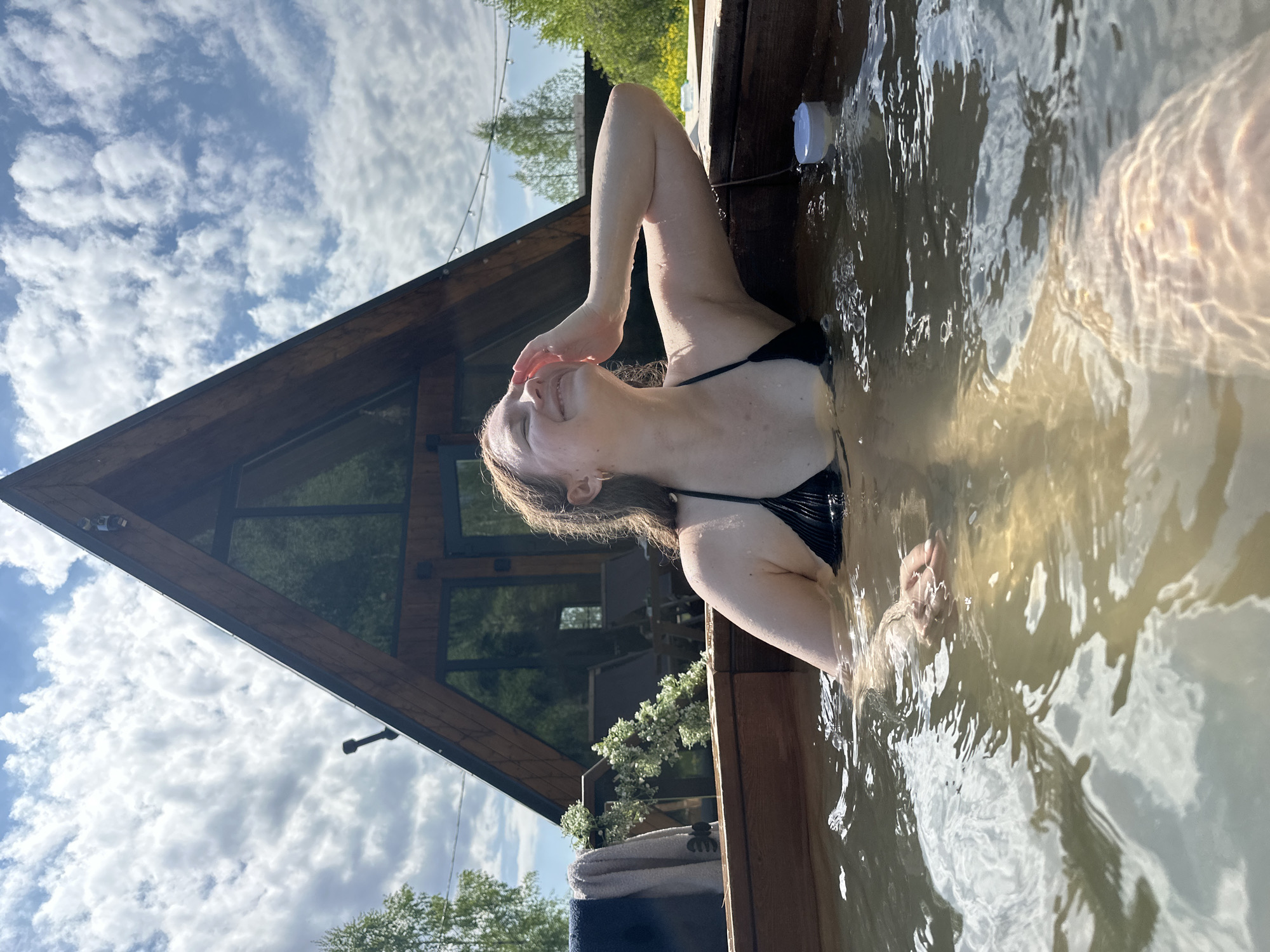 The image depicts a woman in a black bikini in a wooden hot tub. She is leaning against the side of the tub with her right arm raised and her hand touching her forehead. The water in the tub appears murky. Outside the tub, there is a wooden structure with a triangular roof, possibly a sauna or a small cabin. The sky is visible behind the structure and features scattered clouds. The environment suggests an outdoor setting, potentially a backyard or a rural area. While the image does not explicitly confirm the location, it is stated to be Малеевка, Russia.