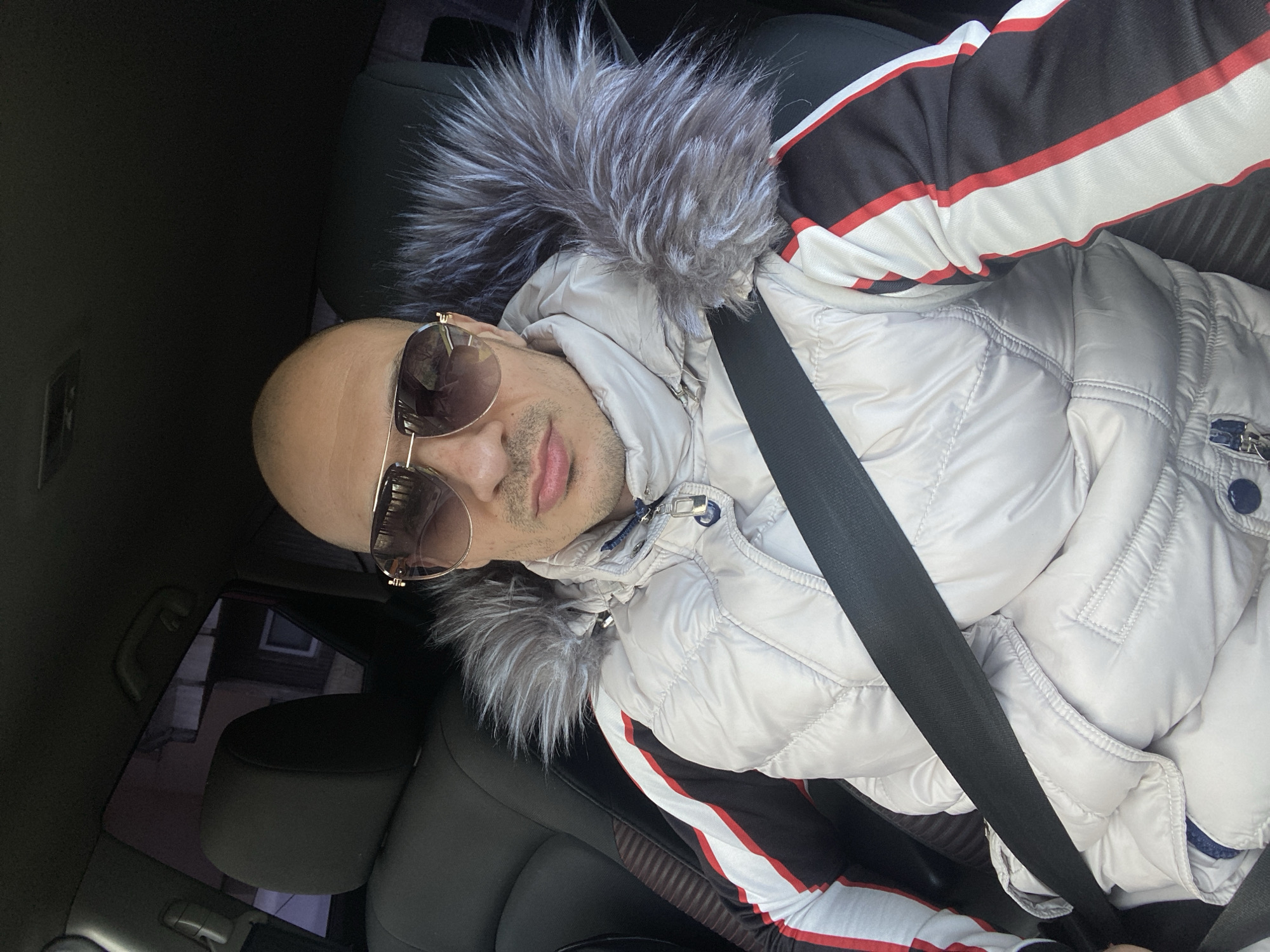 An image depicts an adult male in what appears to be the front passenger seat of a vehicle located in the city of Pleven, Bulgaria. The male, exhibiting a bald head and light skin tone, wears aviator-style sunglasses with gold-colored frames and dark, reflective lenses, obscuring his eyes. Faint facial hair is present on his upper lip and chin. He is dressed in a light grey or silver-toned quilted puffer jacket, which features a prominent grey and white faux fur trim along the hood. Underneath this jacket, a garment with black, white, and red vertical stripes is visible on his right arm. A black seatbelt is securely fastened across his chest and lap. The vehicle's interior is dark, with visible black headrests and seat upholstery. Through the vehicle's left side window, a blurred urban background with indistinct architectural features of buildings is discernible.