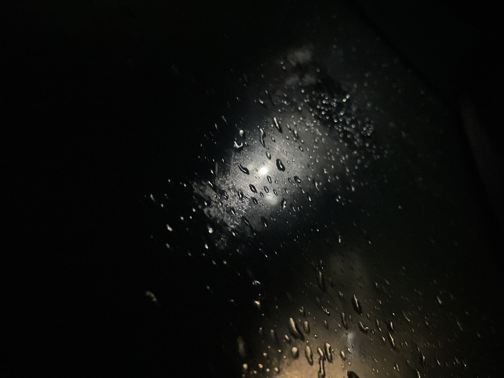 The image shows a dark, rainy scene likely viewed through a window. Water droplets are scattered across the glass surface, refracting light from an indistinct source. The background is mostly black, suggesting nighttime or a very dark environment. The location in Debelt, Bulgaria is not evident from the visual content alone; this detail would require external information or prior knowledge.