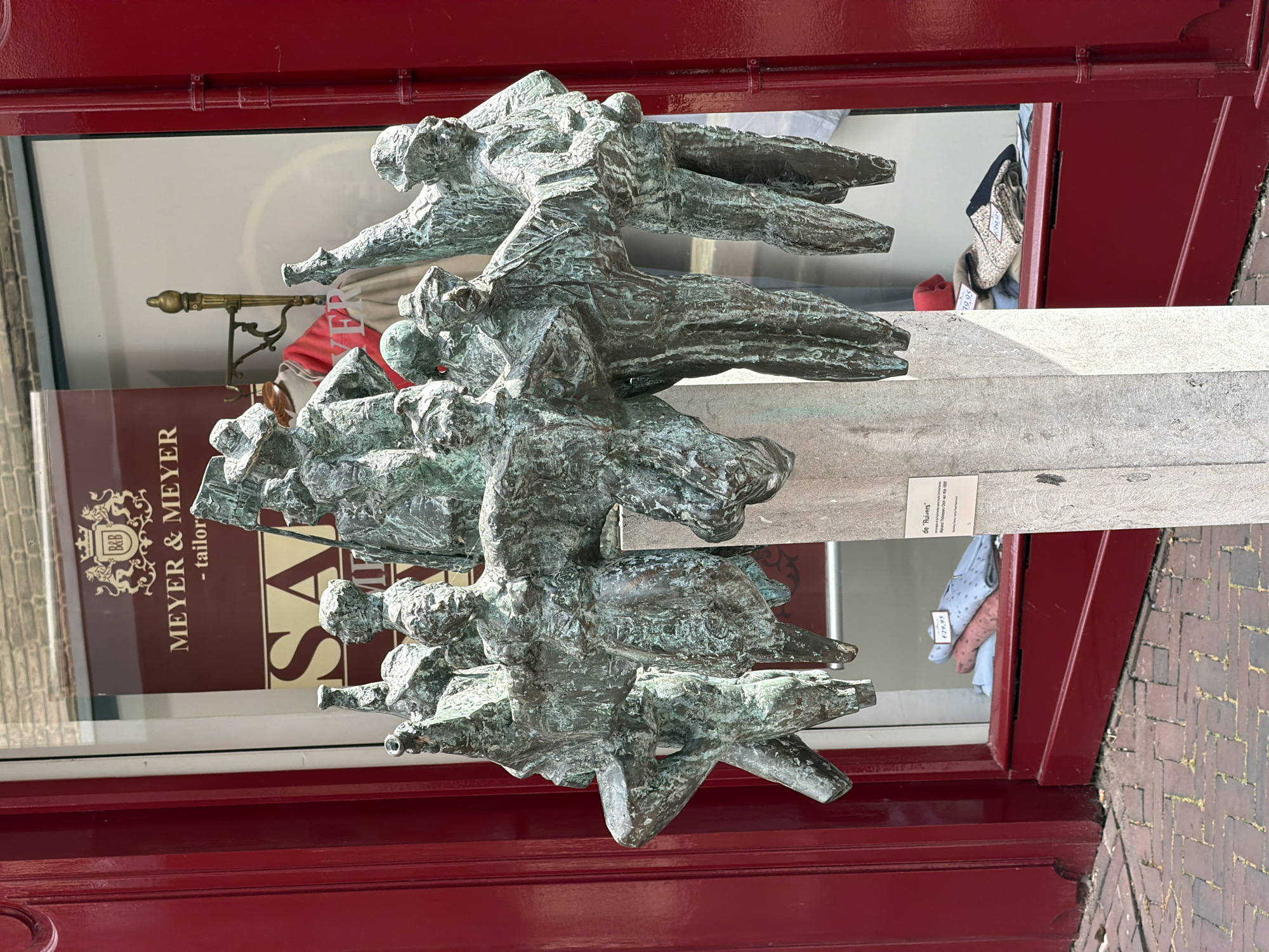This is a medium shot, eye-level, outdoor, eye-level photograph of a bronze sculpture of a group of people on a concrete pedestal, located in front of a building with a storefront in Roermond, Netherlands. The sculpture, which depicts a cluster of figures with abstracted features and stylized clothing, appears to be in motion, with some figures raising their arms and others seemingly marching or dancing. The bronze has a weathered, greenish patina, suggesting age and exposure to the elements.
The building behind the sculpture has a large glass window displaying clothing, with visible price tags. A sign on the window reads "MEYER & MEYR - tailors," and another sign below it says "SALE". The storefront is painted a vibrant red. The sidewalk in front of the building is paved with red brick.
A small, laminated plaque is attached to the concrete pedestal. It is partially obscured by the sculpture but appears to have text written in Dutch. Through the glass, some clothing items are visible in the storefront window, including folded shirts with patterns and some sweaters. The general impression is that of a public art installation in a commercial area. The sculpture's name is "De Ruivers" and it was created by Theo van de Logt in 1992.