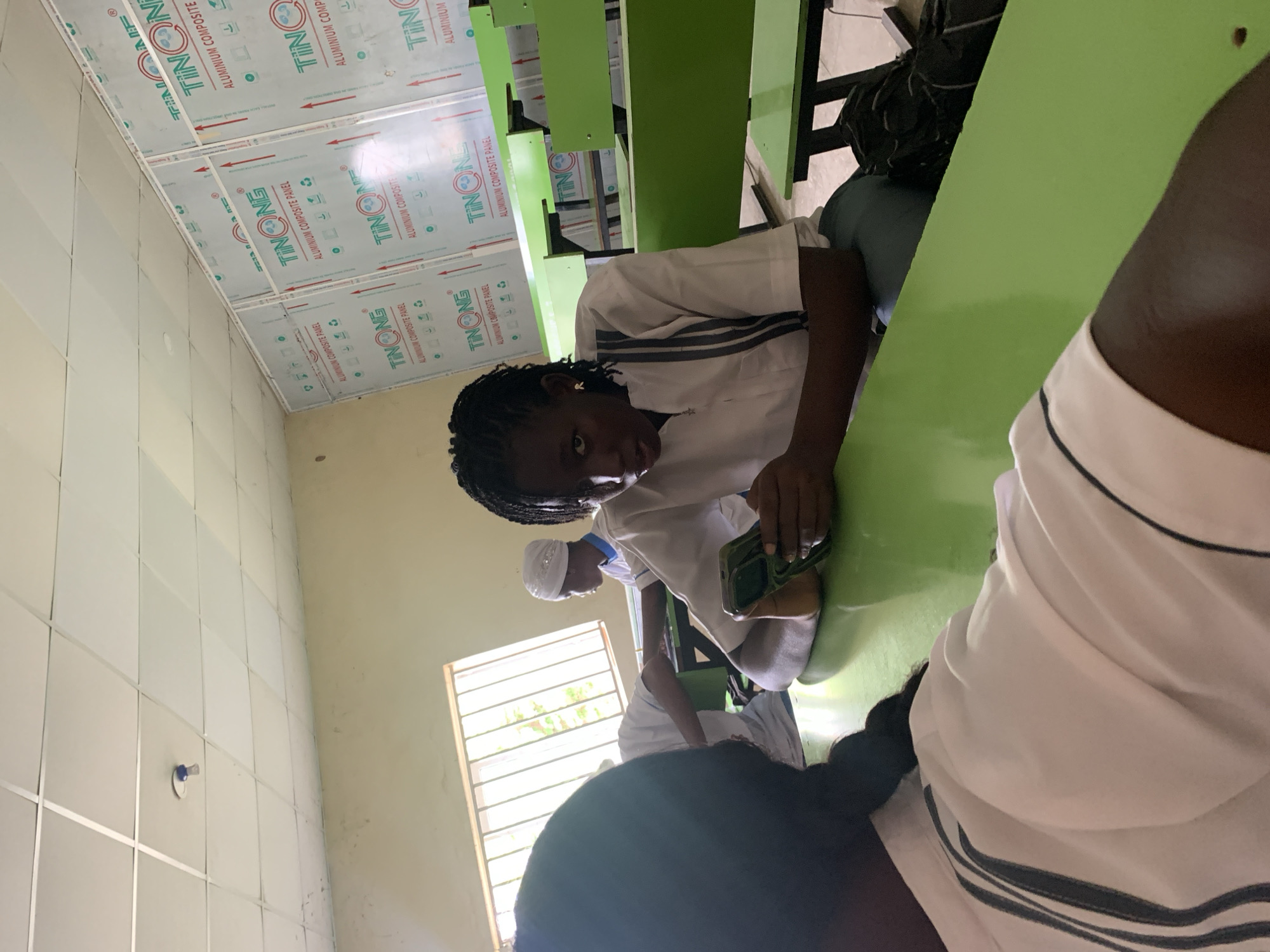 The image shows an indoor scene in what appears to be a school cafeteria or dining hall in Keffi, Nigeria. Several students are seated at long, green tables with attached benches. The students are wearing what appears to be school uniforms, consisting of white shirts with striped detailing. One student in the foreground has braids and is looking directly at the camera while holding a smartphone. Other students are visible in the background, including one wearing a white cap. The walls are tiled with light-colored tiles and sections of the ceiling are covered with aluminum composite panels bearing the "TINONG" brand name. A barred window is visible, letting in natural light.