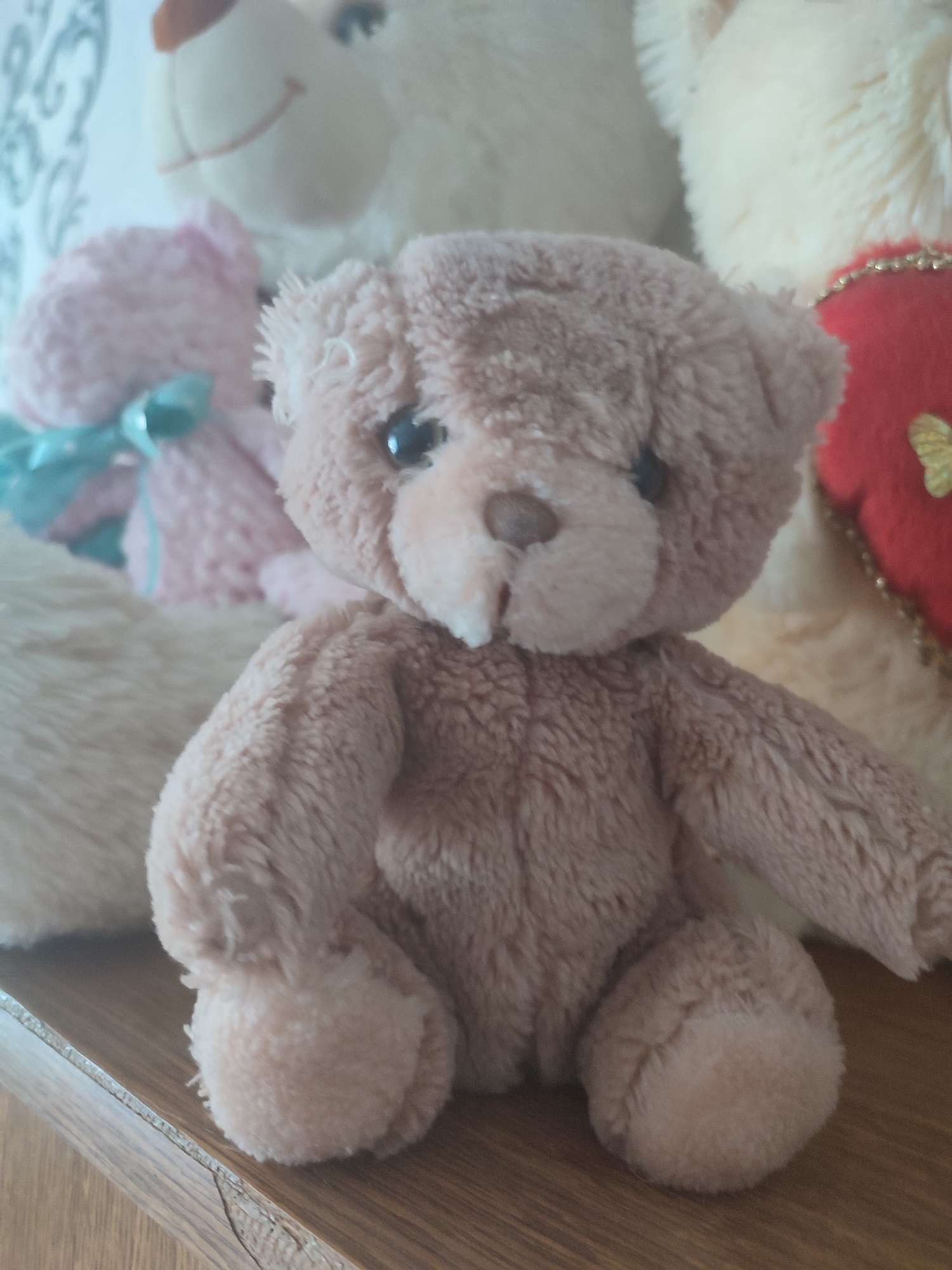 A group of plush teddy bears are arranged on a wooden surface. In the foreground, a small, light brown teddy bear is seated facing the camera. It has a round head with black eyes, a small nose, and a stitched mouth. The bear's body is fluffy, and its arms and legs are positioned slightly outwards. Behind it, to the left, a larger pink teddy bear with a teal ribbon is visible, and further back, a cream-colored teddy bear with a red heart ornament is partially seen on the right. The background consists of a light-colored wall with a subtle pattern. The scene appears to be an indoor setting, possibly a shelf or table.