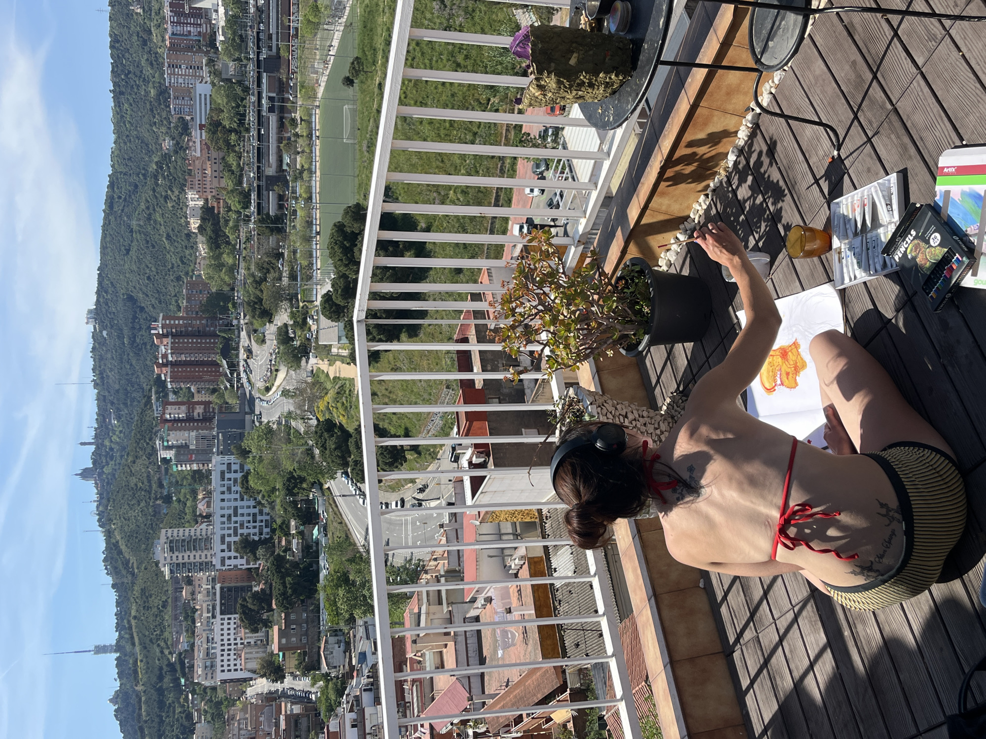A person is painting on a balcony with a panoramic cityscape visible in the background. The person, wearing a red bikini top and striped shorts, is seated on the wooden floor of the balcony, facing away from the camera. They have a paintbrush in their right hand and appear to be working on a painting in front of them. On the balcony, there are several potted plants, painting supplies including gouache, water color pencils, an Artix art set, a glass of liquid, and a small table with a container on top. The balcony has a white railing that offers a view of the city. In the background, a dense urban landscape with buildings of varying heights stretches out towards a green hillside. A soccer stadium is visible. The location is Barcelona, Spain.