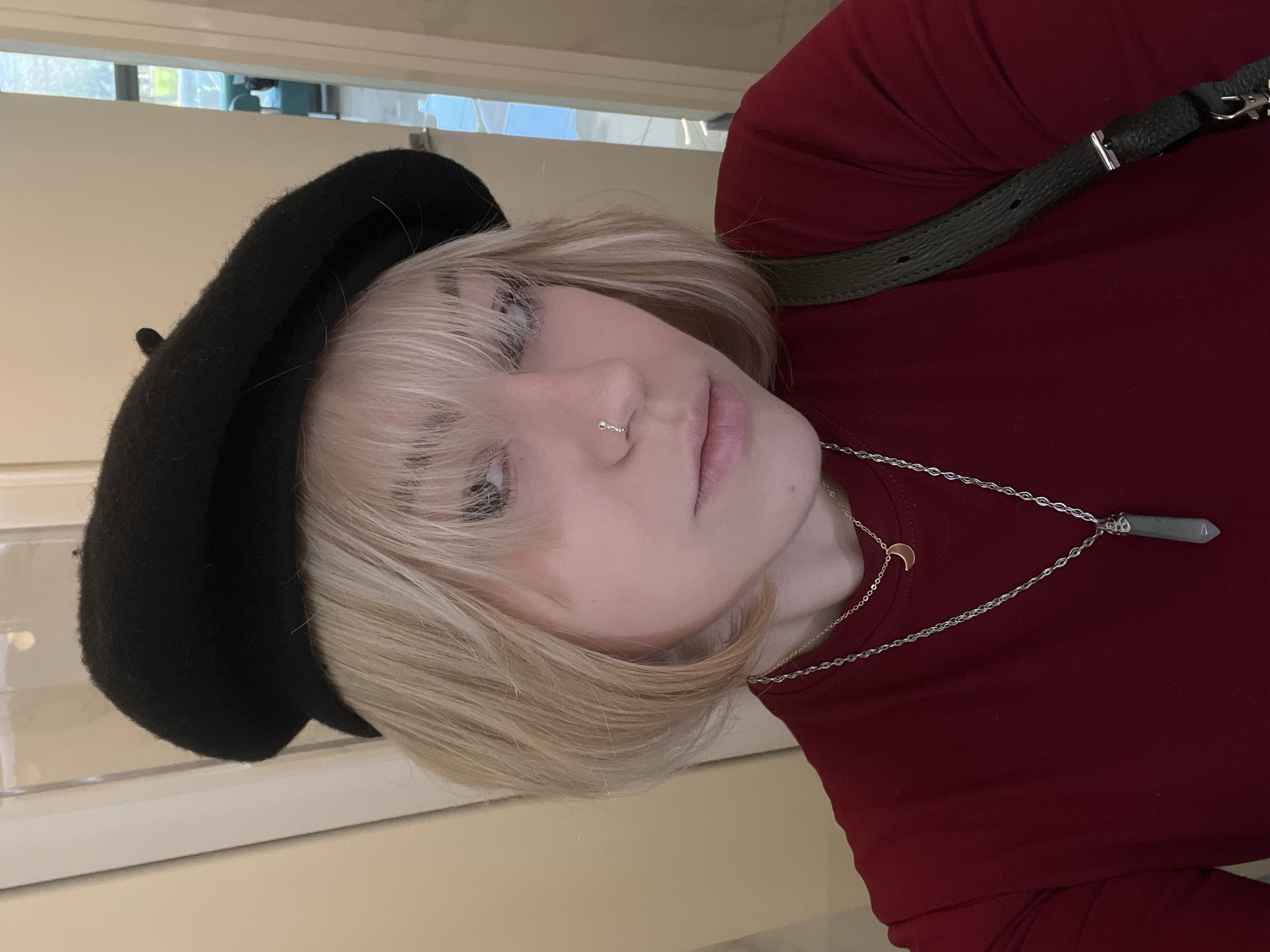 A person with blonde hair, wearing a black beret, a red top, and two necklaces, one gold and one silver with a pointed pendant, is facing the camera. They have a silver nose ring in their left nostril. The background is a blurred interior space with what appears to be a window at the top. The photo is taken at a tilted angle. There is no evidence to suggest this photo was taken in the city of Odesa, Ukraine.