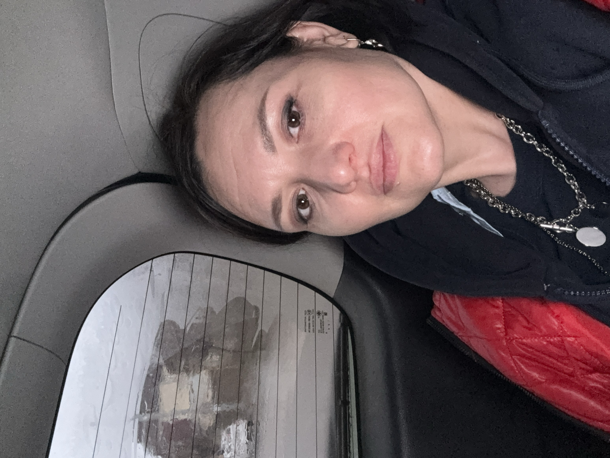 A woman is reclining against a car seat, her head resting on the headrest. The interior of the car is visible, including a rear window with visible vertical lines, potentially a defroster grid. A quilted red jacket or garment is partially visible near her shoulder. The woman is wearing a dark jacket, and a silver-colored necklace is visible around her neck. Her facial expression appears neutral. The location cannot be determined from the image alone. It cannot be placed within the city of Derbent, Russia.