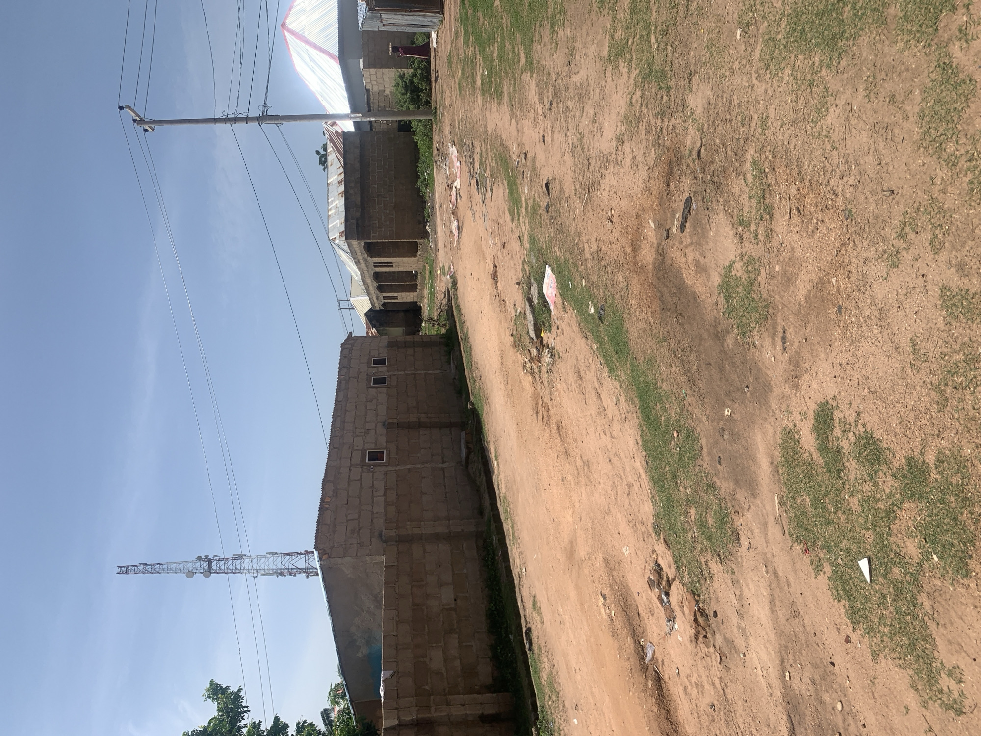 The image depicts a dirt road in Jauro Yinu, Nigeria. A tall telecommunications tower is visible in the distance, along with power lines. Several low-rise buildings constructed with concrete blocks and corrugated metal roofs are situated along the road. The ground is a mix of dry, reddish-brown dirt and patches of sparse green grass. There is some litter scattered on the road and its verges. The sky is mostly clear blue with a few scattered clouds. The overall scene suggests a semi-urban or rural environment under a bright, sunny sky.