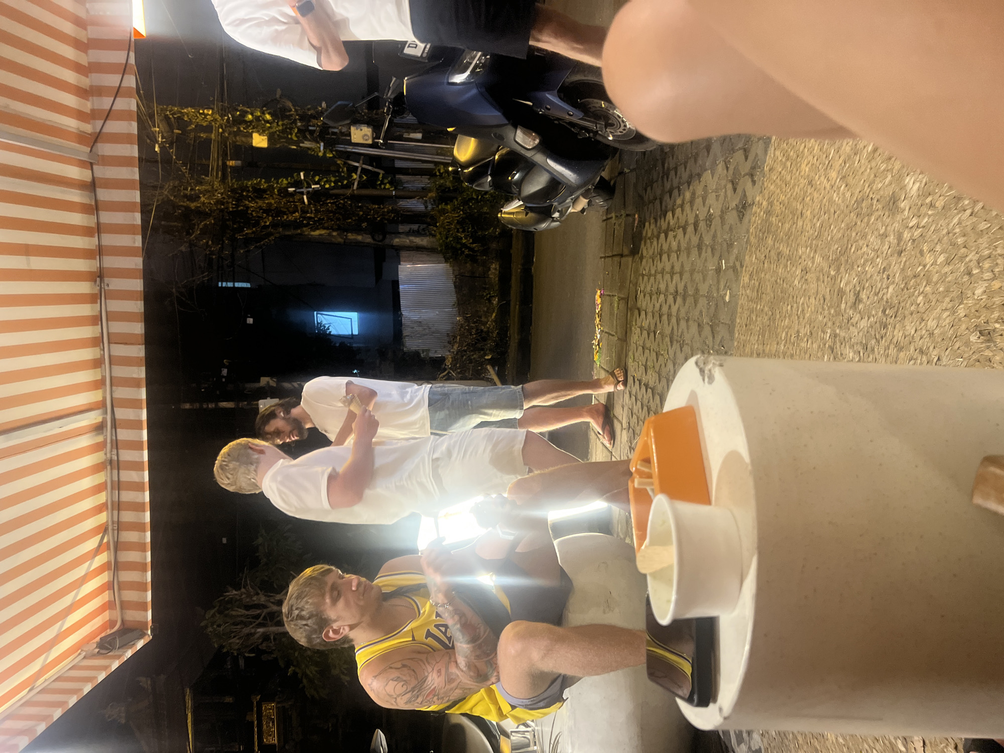 The image captures a nighttime street scene in North Kuta, Indonesia, with four men gathered around what appears to be a street vendor's stall. The setting is illuminated by artificial lights, creating a warm glow around the subjects against the dark background.
In the foreground, a man with tattooed arms is seated, wearing a yellow basketball jersey with "Lakers" and the number "12" clearly visible. He is looking towards the right, seemingly engaged with an unseen person or activity. Directly in front of him is a large, white cylindrical container, likely part of a vendor's setup, holding white cups and wooden spoons, suggesting a stall selling beverages or ice cream.
Three other men are standing near the stall. Two are wearing white t-shirts and shorts; one is in lighter shorts and flip-flops, holding a small cup or container. The third standing man is partially visible to the left, also in a white t-shirt. The group appears to be casually socializing or waiting for service from the vendor.
In the mid-ground and background, multiple scooters are parked, a characteristic feature of Indonesian streets. To the far left, a distinct orange and white striped awning extends over the scene. The ground is paved, and some dark vegetation is visible further back, adding to the outdoor, street-side ambiance. The overall impression is a relaxed, informal gathering at a food or drink stall on a warm evening.