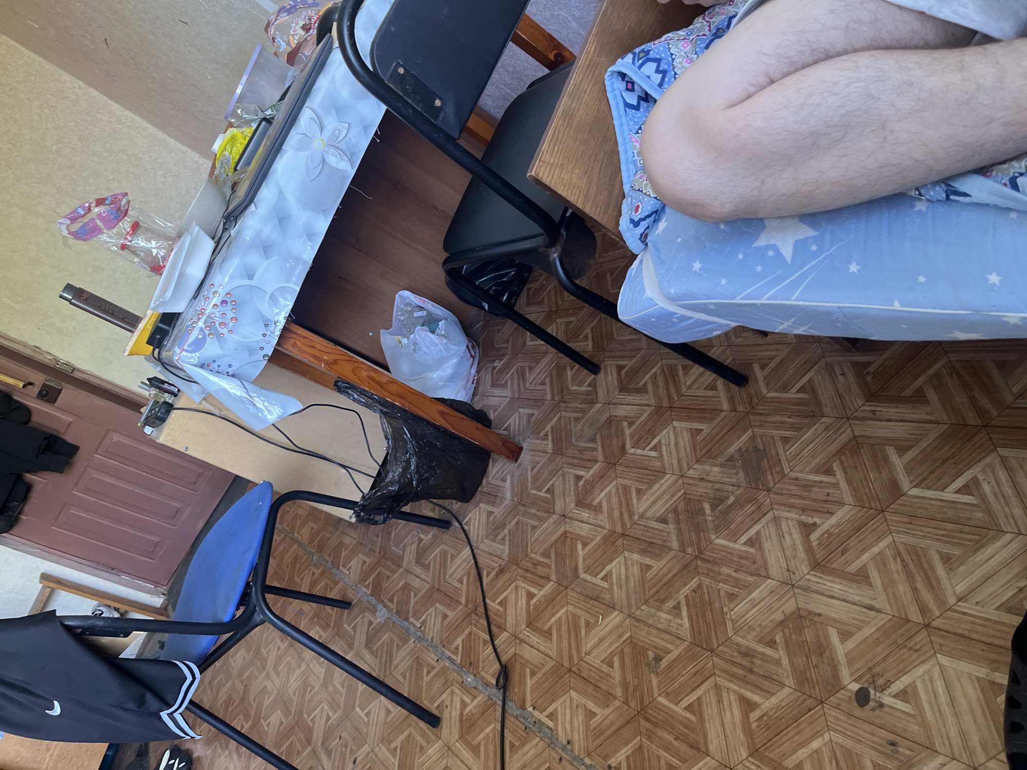The image depicts an indoor scene that appears to be a room or living space, possibly in an apartment or house. The flooring is made of wooden tiles with a geometric pattern in shades of brown and beige.
In the foreground, a person's legs are visible, resting on a blue mattress with a white star pattern. The legs are fair-skinned with visible hair.
To the left, a black metal chair with a blue seat is positioned. A pair of black athletic shorts with white stripes is draped over the back of the chair.
Further back, a table is covered with a light-colored tablecloth featuring a floral design. On the table are various items, including a bottle of what appears to be shampoo or conditioner, a white plastic container, and several plastic bags containing objects. A black laptop or similar electronic device is also on the table.
Behind the table, there is a wooden door, likely an interior door, painted a reddish-brown color. To the right of the door, a part of a wall is visible, painted a light beige color.
In the middle ground, a black plastic trash bag is situated on the floor, partially obscuring a black chair. Another white plastic bag, likely containing trash or discarded items, is near the black chair. A black electrical cord runs across the floor.
The overall impression is one of a lived-in space, possibly slightly cluttered, with everyday objects present. The scene does not offer specific clues about the exact location within Krasnye Baki, Russia, beyond being an indoor residential setting.