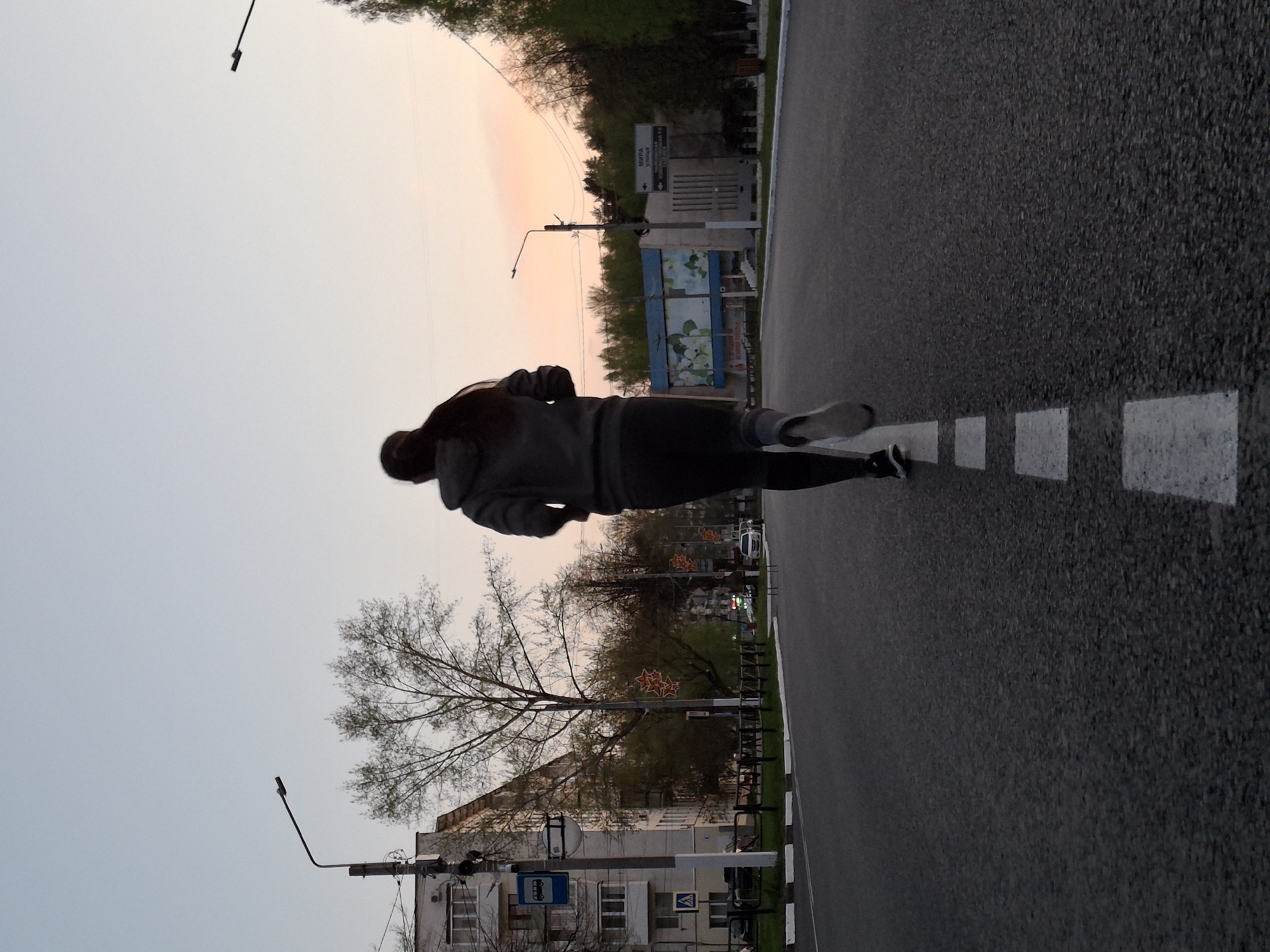 A young person is jogging on Mira Street in Chaikovsky, Russia, facing the direction of the intersection of Mira Street and Sovietskaya Street. The individual is wearing dark pants and a grey hooded jacket. The road has white dashed lines indicating a traffic lane. In the background are buildings, street lights, trees, and decorations of red stars on some trees. The sky is light grey, likely near dusk.