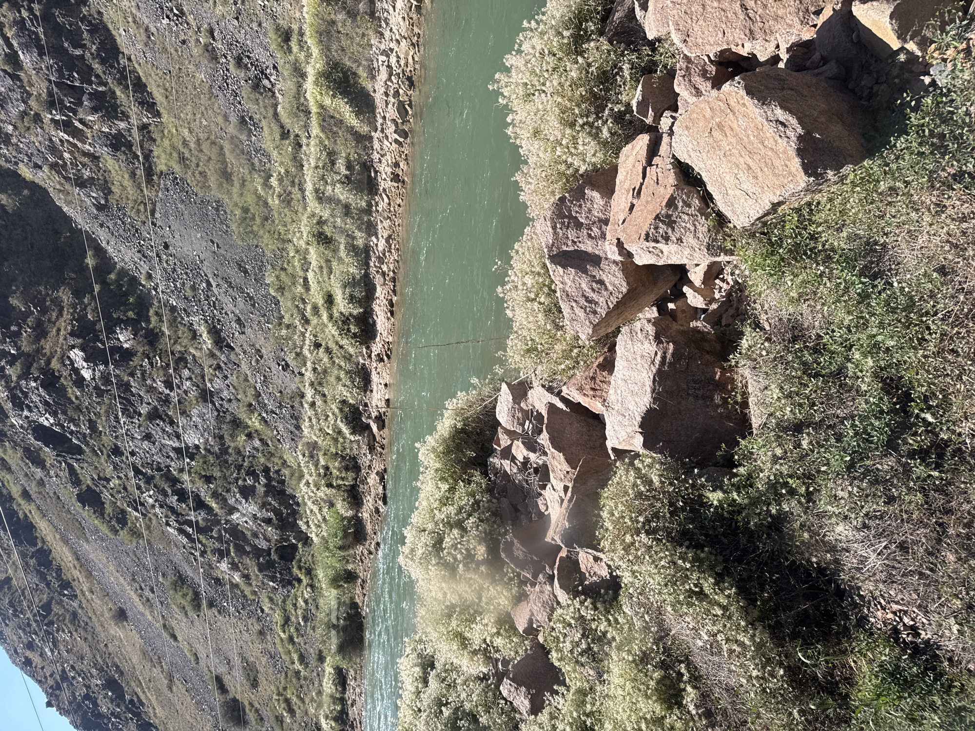 The image shows a river flowing through a mountainous landscape. The river is a vibrant green color and appears to be flowing swiftly. The banks of the river are lined with rocks and vegetation. The mountains in the background are steep and rocky, with sparse vegetation. There are power lines running across the mountainside. The scene is brightly lit by the sun, casting shadows on the rocks and vegetation. The location is identified as Krasnyy Most, Kyrgyzstan.