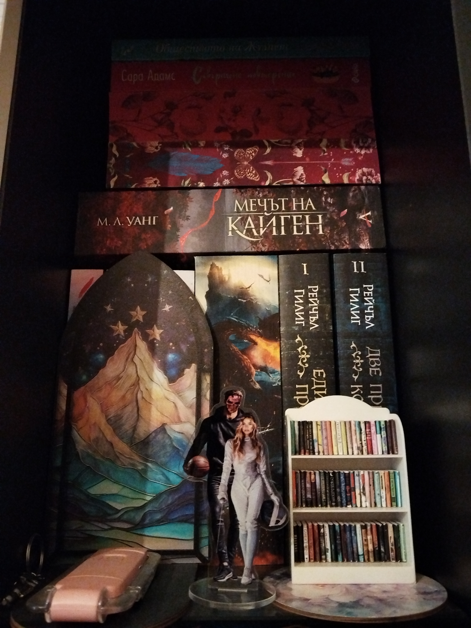 The image displays a close-up of a dark bookshelf or display shelf, arranged with books and decorative objects.
On the top section, three hardcover books are stacked horizontally. The topmost book has a dark teal spine with partially visible Cyrillic text reading "Общественото на..." (The Public of...). Below it, a red book features a spine with ornate floral patterns and Cyrillic text identifying "Сара Адамс Сборник..." (Sara Adams Collection...). The third book, also red, exhibits similar floral motifs alongside butterflies and an arrow design.
Below these, a larger, dark-covered book is positioned horizontally. Its cover features a muted, dark forest or nature scene, with Cyrillic text clearly visible: "М. Л. УАНГ" (M. L. Wang) on the left, and "МЕЧЪТ НА КАЙГЕН" (The Sword of Kaigen) on the right.
In the foreground, standing upright, are several items. On the far left, a dark, arch-shaped decorative piece functions as a book stand, depicting a stylized mountain range under a dark, starry sky with four prominent golden stars. To its right, two clear acrylic standees of human figures are positioned. The male figure, on the left, wears a black hooded top and pants, holding a basketball; his face appears to have reddish scarring or paint. The female figure, on the right, is dressed in a light grey or silver form-fitting suit and holds a helmet in her left hand.
Behind these standees, two dark-spined books are vertically arranged. Both spines display the Cyrillic author name "РЕЙЧЪЛ ГИЛИГ" (Rachel Gillig). The left book is marked "I" followed by partially visible text "ЕДИН ПР...", and the right is marked "II" followed by "ДВЕ ПР...". These likely correspond to volumes I and II of a series.
On the flat surface in front of these items, a small, pink, opaque object, possibly a miniature car or futuristic vehicle, lies horizontally on the left. To its right, a miniature white bookshelf with three shelves is filled with numerous tiny, colorful books. The circular surface directly beneath these items exhibits an abstract, swirling pattern in shades of blue, purple, and brown, resembling polished stone or a painted coaster.
The visible text on all books is in Bulgarian Cyrillic. The specific location in the city of Plovdiv, Bulgaria, cannot be determined from the provided image.