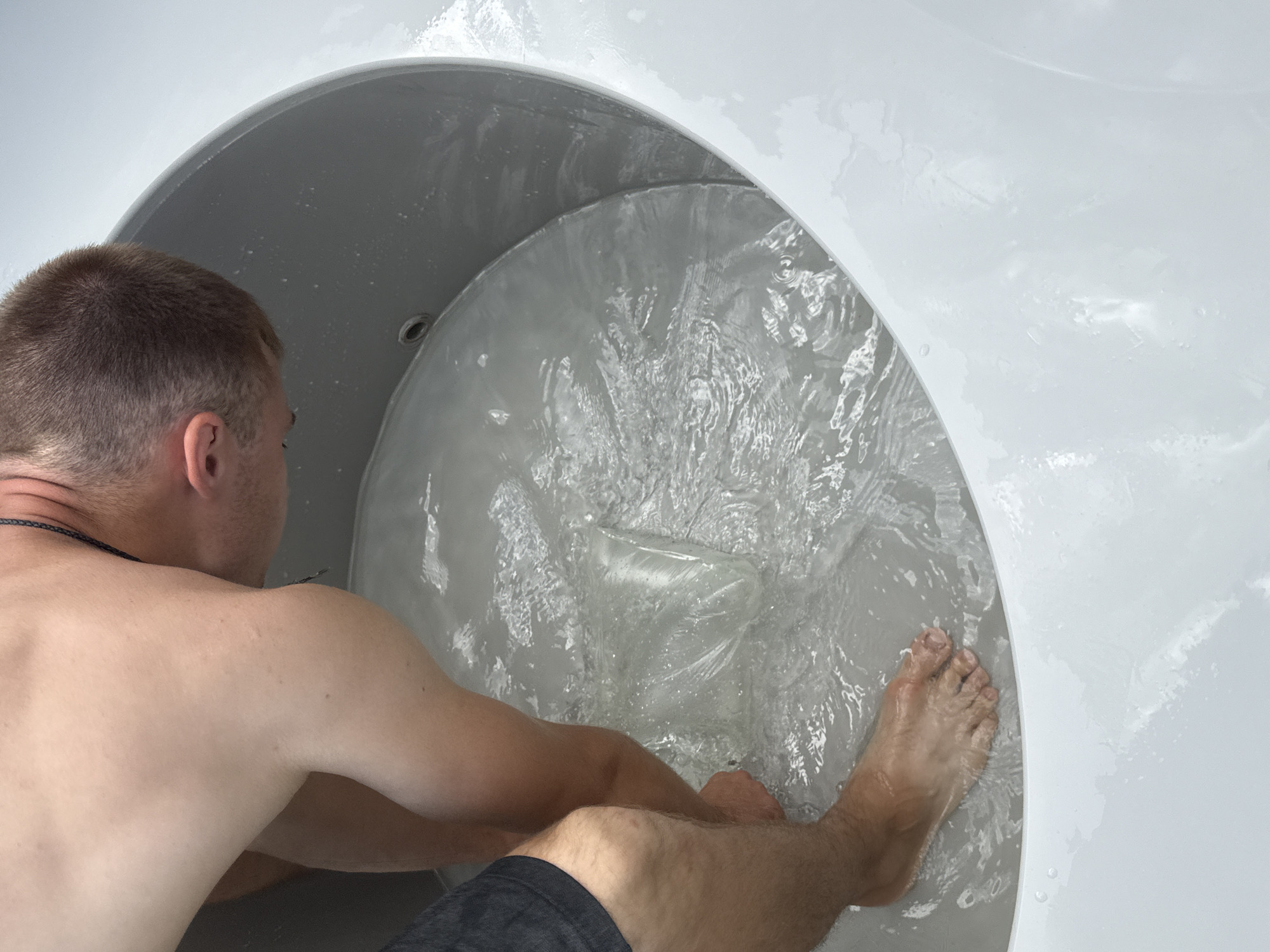 The image shows a person's torso and legs submerged in a round, grey tub filled with clear water. The person is leaning forward, with their head and shoulders obscured from view. One of the person's bare feet is visible in the water, as is a clear plastic bag containing an unknown substance. The tub appears to be made of plastic and is partially filled with water, creating ripples and reflections on its surface. The background is out of focus, but suggests an outdoor setting. The image is taken from a high-angle perspective, looking down into the tub. The image does not contain any identifiable landmarks or elements that would indicate a specific location in Starokorsunskaya, Russia.