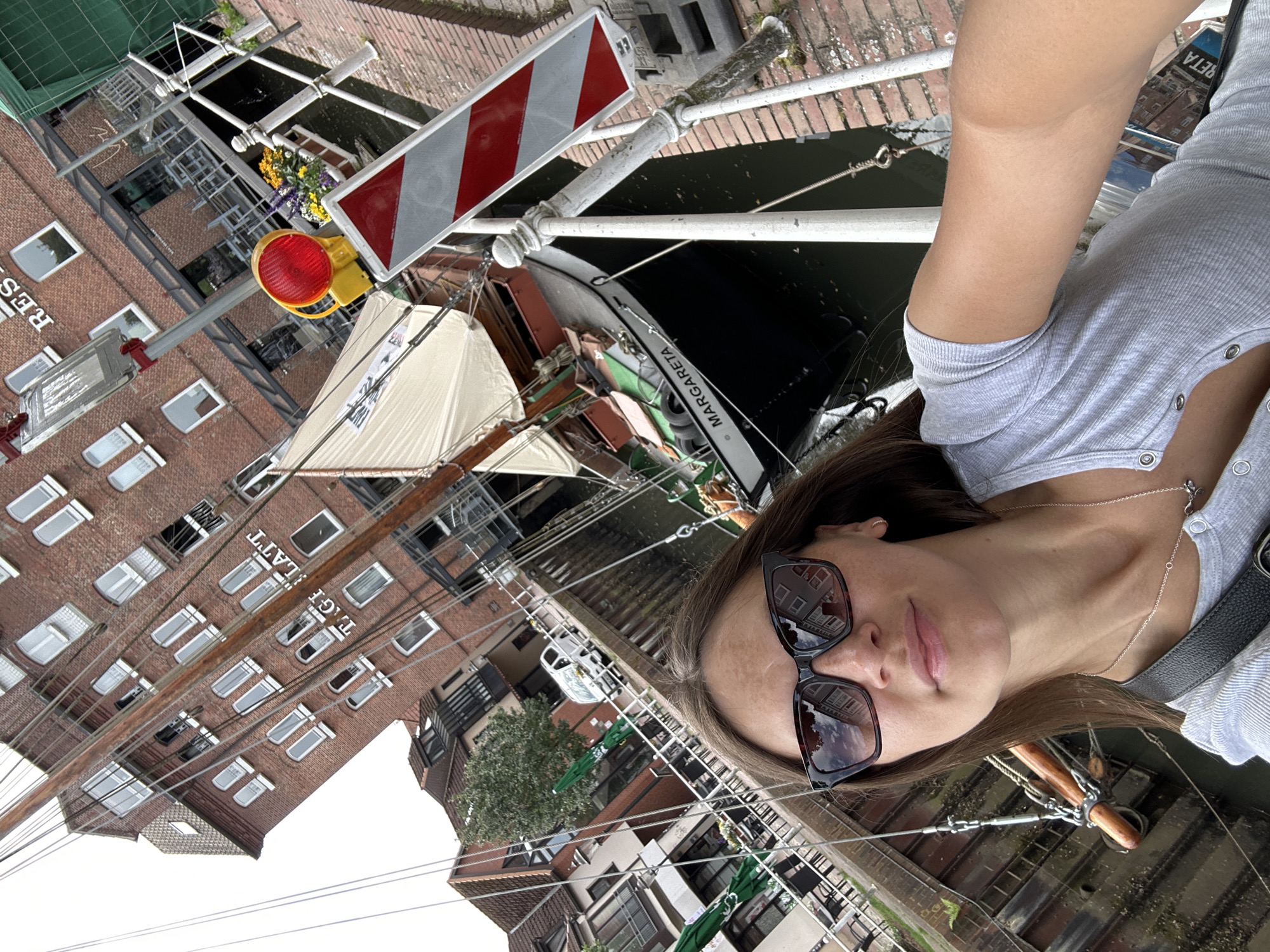 A person is taking a selfie on a bridge in Buxtehude, Germany, overlooking the canal. The shot includes portions of the person's face and torso. The bridge is lined with white railings. A boat named "Margareta" is visible in the canal. A building with a red brick facade and the name "Tageblatt" is on the left side of the image. Traffic control signals and signs are attached to the bridge structure.