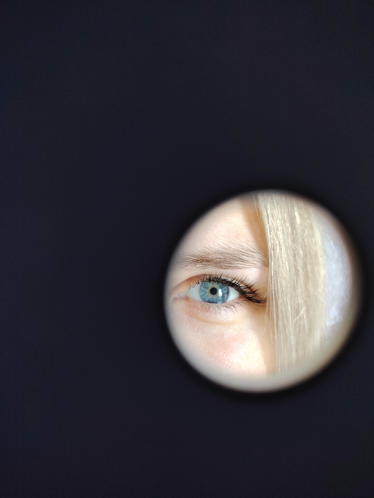 A close-up shot shows a person's blue eye and part of their face, framed within a circular opening against a dark background. Strands of blonde hair fall across the right side of the face. The location in the city of Chaikovsky, Russia is not discernable from this image.