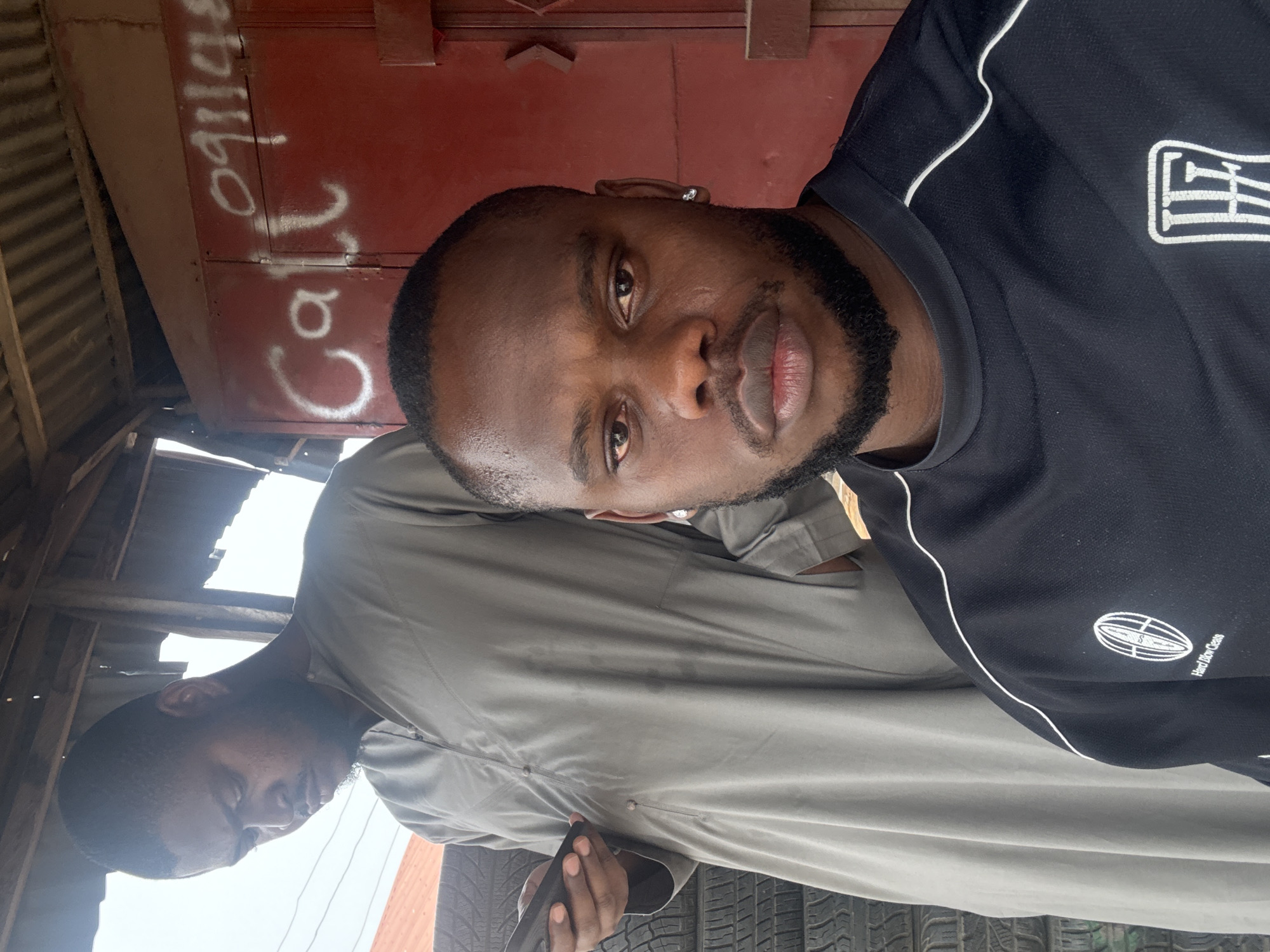 A portrait-style photograph depicts two Black men in what appears to be an outdoor commercial setting in Lugbe, Nigeria, under bright daylight conditions.
The foreground features a young man with a short, neat haircut and a well-trimmed beard, looking directly at the camera with a neutral and somewhat serious expression. He is wearing a black short-sleeved athletic shirt with white piping along the shoulders and sleeves. On the right chest of his shirt, a white oval logo with internal horizontal lines is visible, beneath which the text "Hard Row Gone" can be read. He has a silver stud earring in his left ear.
Partially obscured behind him and to his left stands another man, dressed in a light grey long-sleeved garment, possibly a traditional tunic. His face is mostly in shadow, and he appears to be looking downwards or away, holding a black smartphone in his hands.
The background consists of a reddish-brown metallic surface, likely a shop door or shutter, on which white graffiti is scrawled, reading "Call 09114s" (the last character is slightly ambiguous, appearing as 's' or '5'). Above this surface and to the left, a corrugated metal roof structure supported by wooden beams is visible, suggesting a sheltered stall or small business. Stacked tires are visible at the bottom of the frame, indicating the location might be a tire repair shop or an automotive service point. The setting conveys an informal, everyday street-side commercial environment.