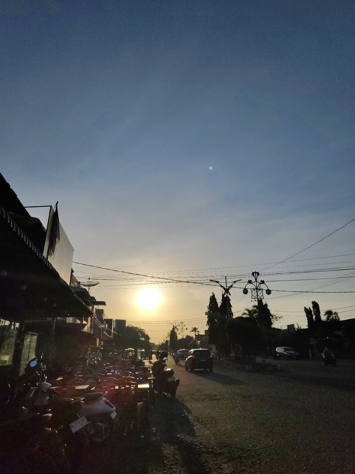 The image depicts a street scene in Lhokseumawe, Indonesia, at sunset. The sun is positioned in the center of the frame, partially obscured by buildings and trees. The sky is pale blue with wispy clouds. On the left side of the street, there are commercial buildings with awnings, and a row of parked motorcycles. Tables and chairs are set out in front of the buildings. On the right side of the street, there are trees and parked cars. Vehicles are driving on the road. Power lines stretch across the sky above the street. There is a bright moon visible. The image is taken at ground level, and the road surface is visible in the foreground.