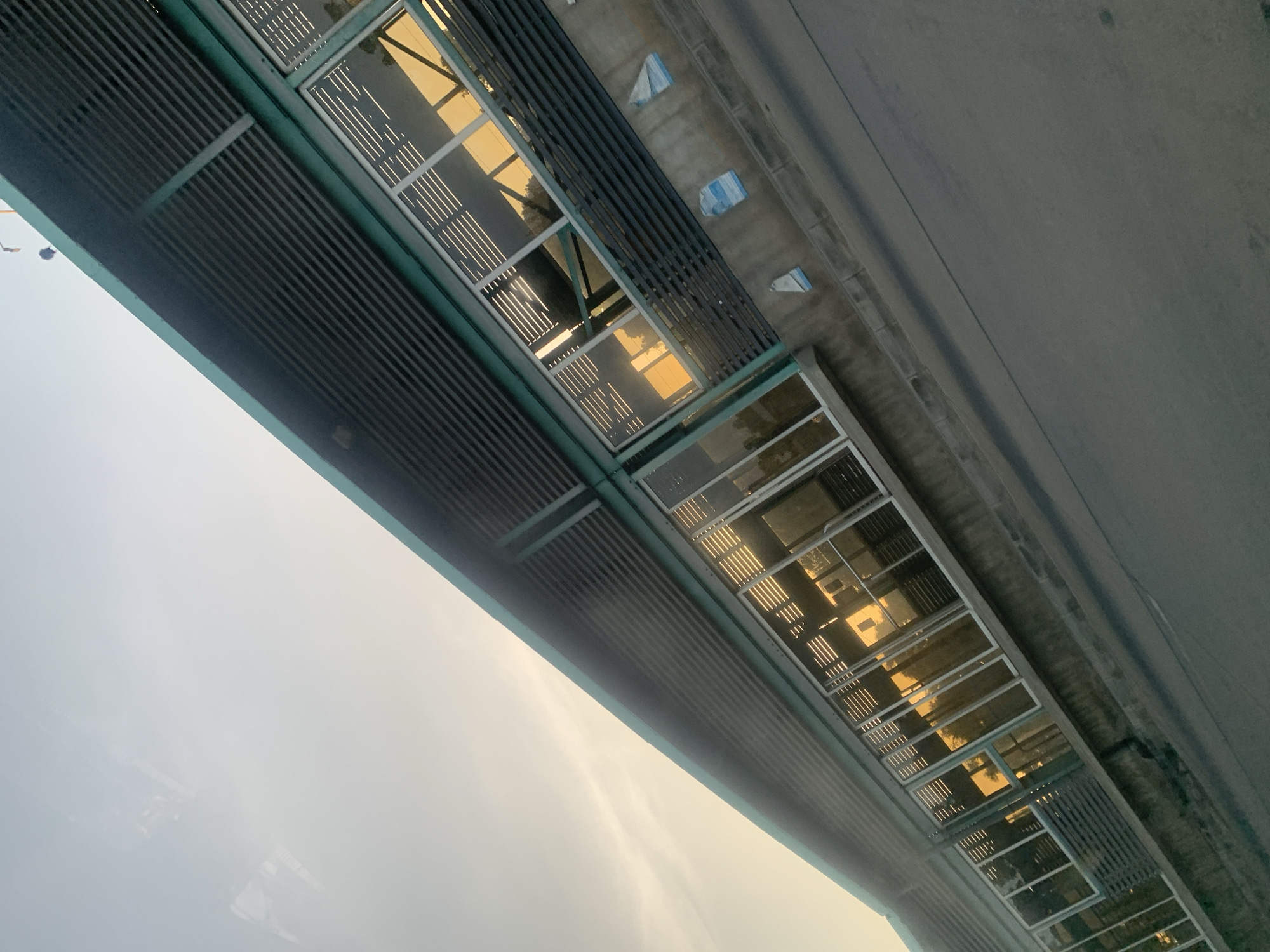 The image shows a section of the Dar es Salaam Bus Rapid Transit (BRT) infrastructure in Tanzania. The BRT system is elevated, with the bottom of the structure visible in the photo. The photograph is taken at a low angle, focusing on the elevated busway. The underside of the road is visible, along with a series of rectangular openings, possibly windows or access points, on the sides of the elevated structure. Above the busway is a wall with regular narrow horizontal slits. The general environment seems to be urban, with reflections of nearby buildings visible in the windows, and the sky is bright and somewhat hazy, suggesting early morning or late afternoon lighting.