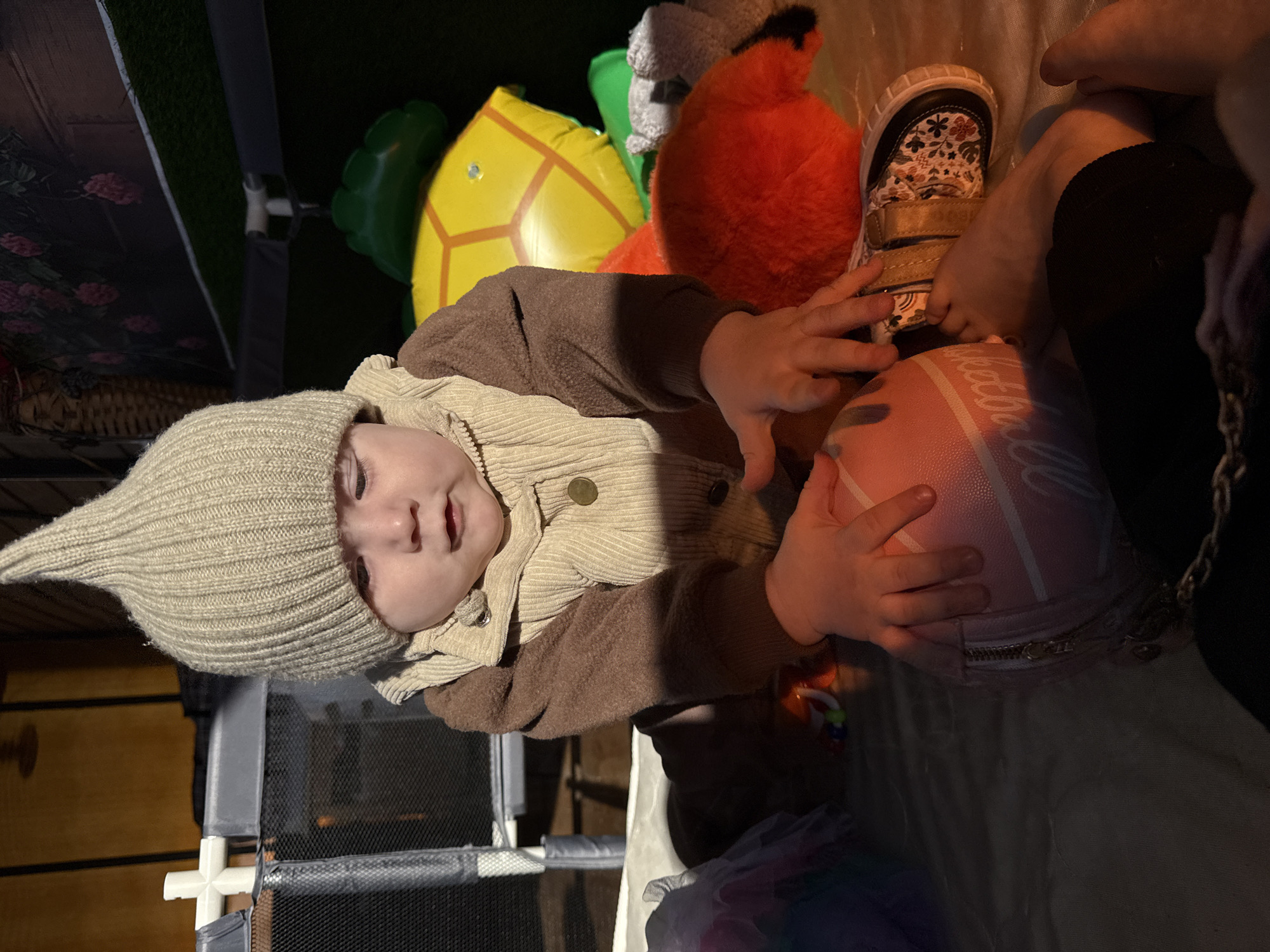A baby in a brown jacket and beige pointed hat plays with a pink purse. The baby is indoors, next to a playpen. In the background are a yellow inflatable turtle and a red plush toy. The image appears to have been taken at night or in a dimly lit location. There is no information in the image to ascertain location in the city of Кипчак, Russia.