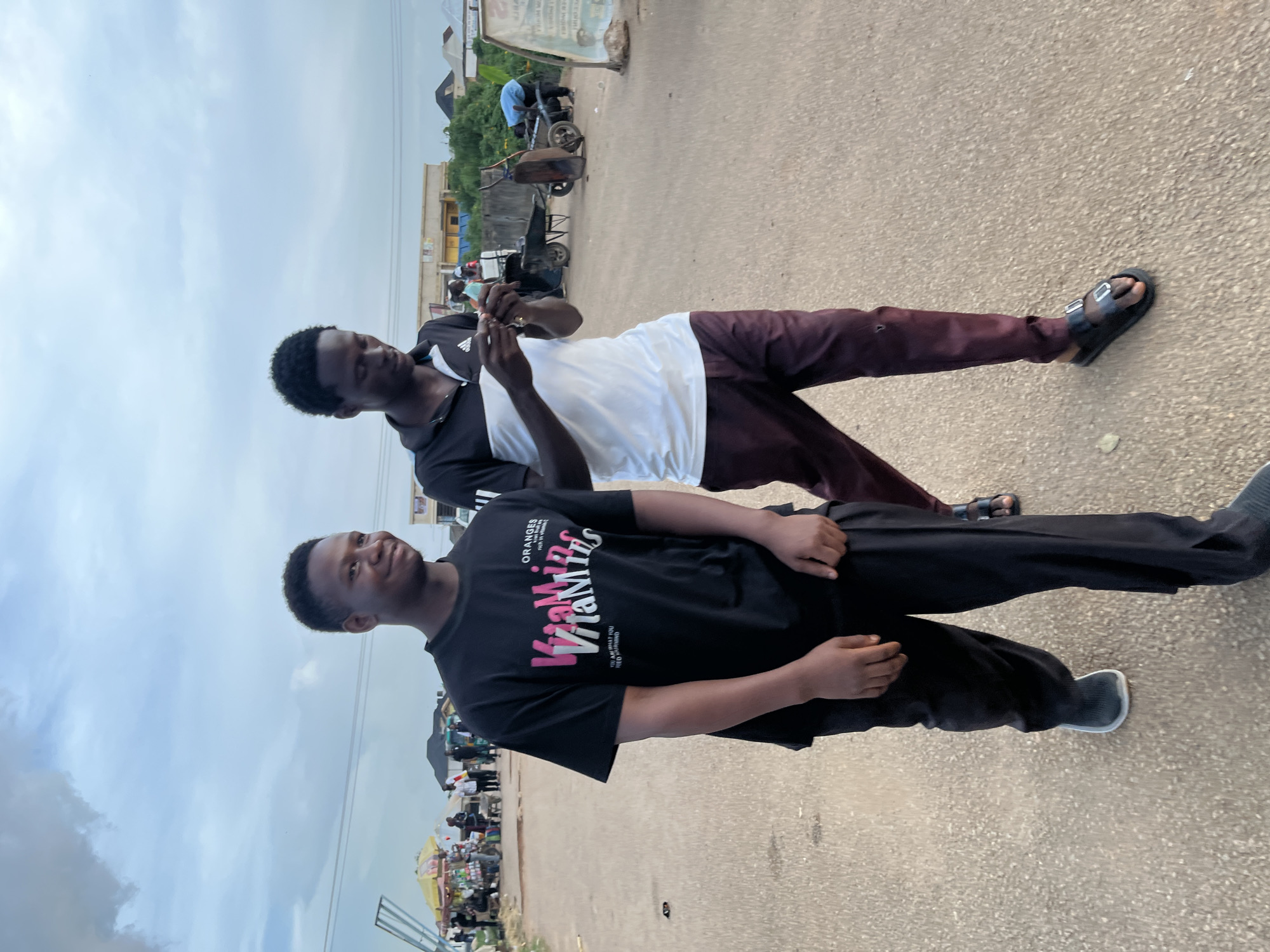 Two young men are standing outdoors on a paved street or pathway in what appears to be Rafin Par, Nigeria. The sky is overcast, indicating it is daytime.
The man in the foreground, positioned on the left, is wearing a black t-shirt, black pants, and dark sneakers with white soles. His t-shirt features prominent text: "ORANGES" at the top, "Viamins" (likely stylized "Vitamins") in pink lettering, and smaller text below that reads "THE NEW WAVE YOU'RE GOING TO EXPERIENCE WE GET INSPIRED." He has short, dark hair and a slight smile, looking generally towards the viewer.
Behind him, on the right, stands another man. He is wearing a two-toned polo shirt, black on the upper half with a white section below, and a small white Adidas logo is visible on his left shoulder. He has dark maroon or brown pants and dark sandals. This man is holding a smartphone with both hands, appearing focused on its screen.
The background shows a lively street environment. On the right, there are various buildings, some with yellowish walls, and what look like market stalls or small shops with goods displayed. Several vehicles, including a tricycle (keke-napep) and a wheelbarrow, along with other people, are visible further in the distance, suggesting a bustling urban or semi-urban setting.
