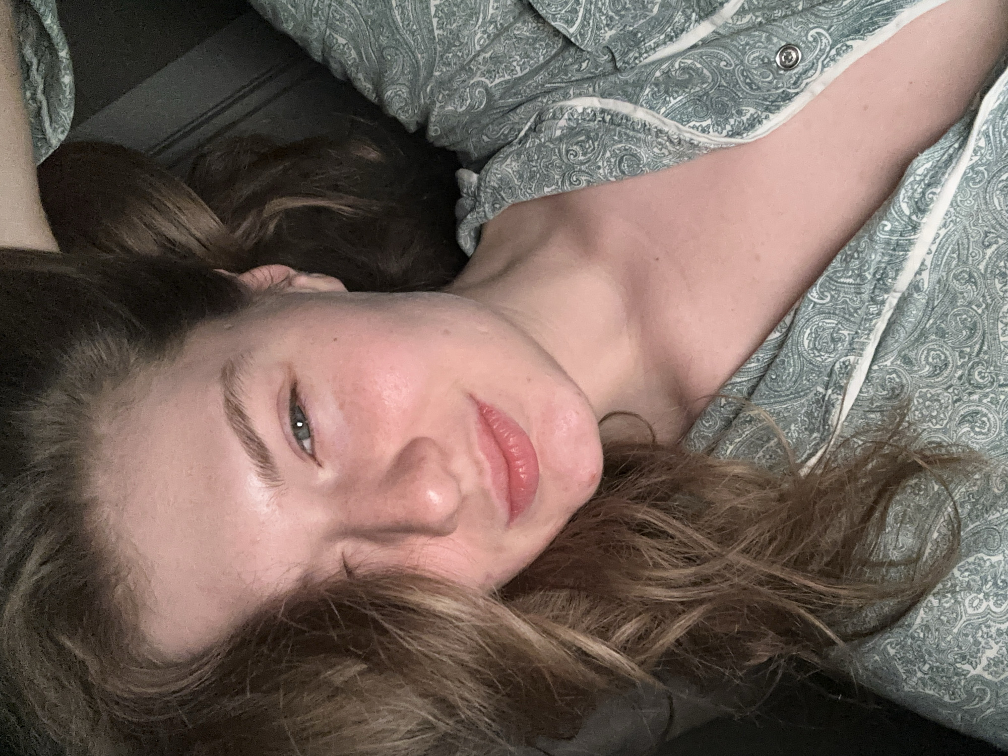 A light-skinned individual with long, light brown hair is lying on a dark surface, possibly a bed or sofa. The person is wearing light green and white paisley patterned pajamas, which are partially bunched around the neck and upper body. The person's face is the primary focus, displaying a subtle smile. The background appears to be dark and indistinct, suggesting an indoor setting with low lighting. The image does not provide any visual elements that could place it within the city of Kazan, Russia.