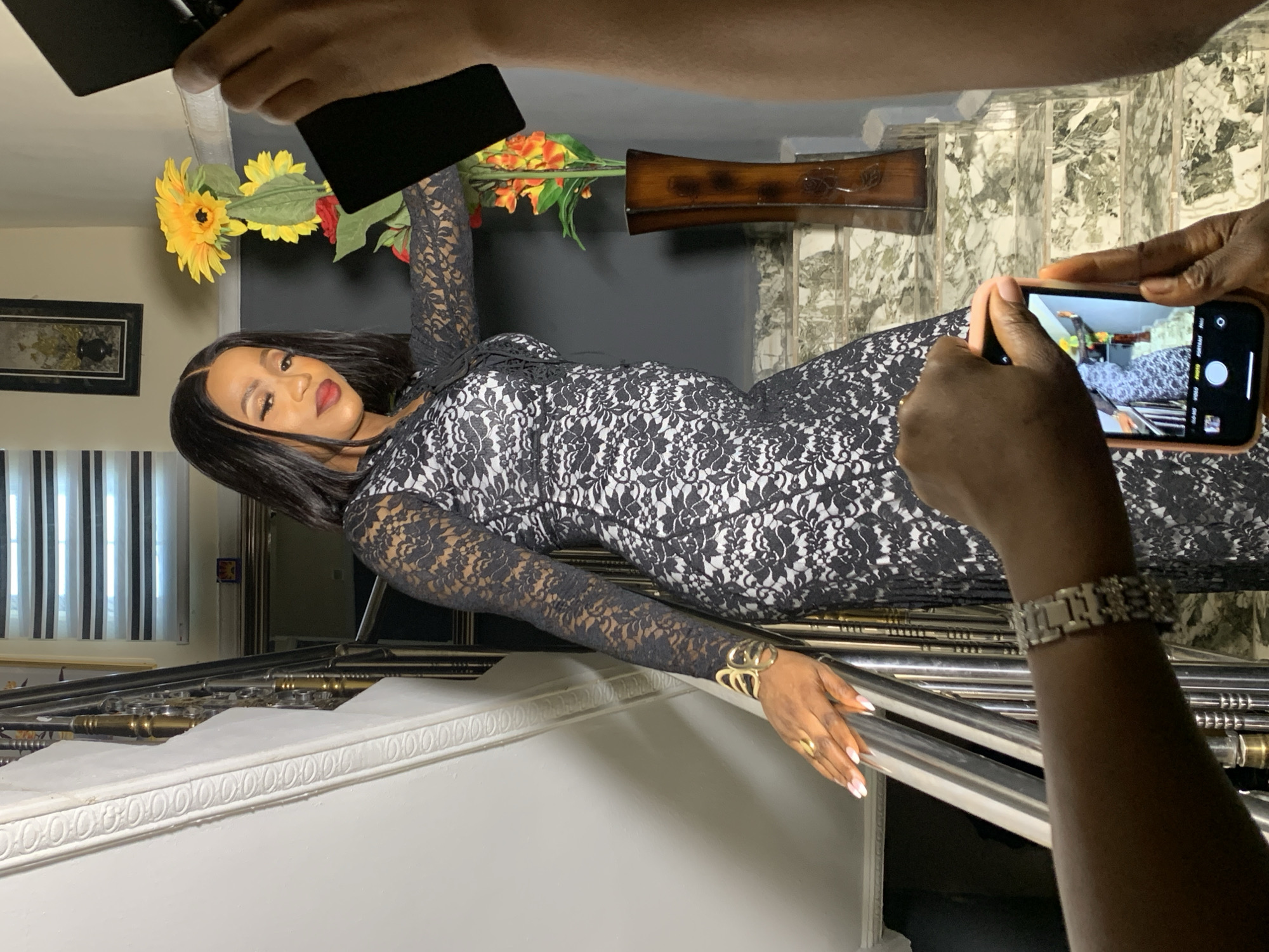 A woman lies on a set of stairs, posing for a photo in what appears to be an indoor location. The woman wears a black and white patterned dress, long-sleeved, and gold jewelry on her wrist and hand. Her dark hair is styled to the side, and she has full makeup. Two individuals are visible taking photos of her, one using a smartphone and the other a device that appears to be a flash or reflector. The location includes a staircase with a metal railing, decorated with what seems to be a flower arrangement behind the woman. A framed artwork hangs on the wall, and blinds are visible on a window. The setting gives the impression of a domestic interior. The city of Jos, Nigeria is not apparent from visual clues in this image.