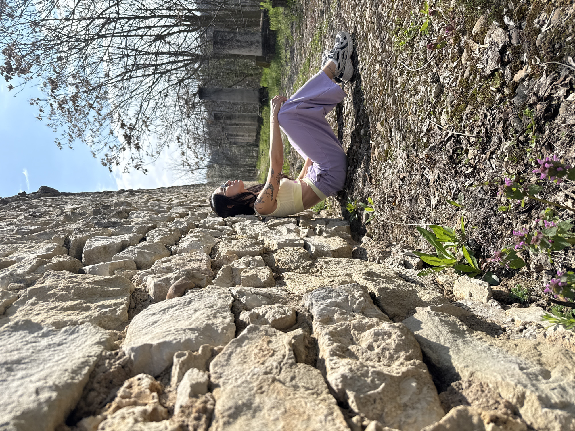 A young woman lies on her back on a stone wall in Pleven, Bulgaria. The wall is made of light-colored, rough-hewn stones of varying sizes. The woman wears a light yellow top, purple pants, and sneakers. She has dark hair and is positioned with her head toward the left of the frame and feet toward the right. Her knees are bent, and her hands are clasped in front of her chest.
Behind the woman, the ground slopes upwards, covered in dirt, leaves, small rocks, and some green foliage with small purple flowers. Faintly visible in the background are bare trees and some indistinct structures or buildings. The sky is blue with scattered clouds visible in the upper left corner. The angle of the photo creates a distorted perspective, making the wall appear steep and uneven.
