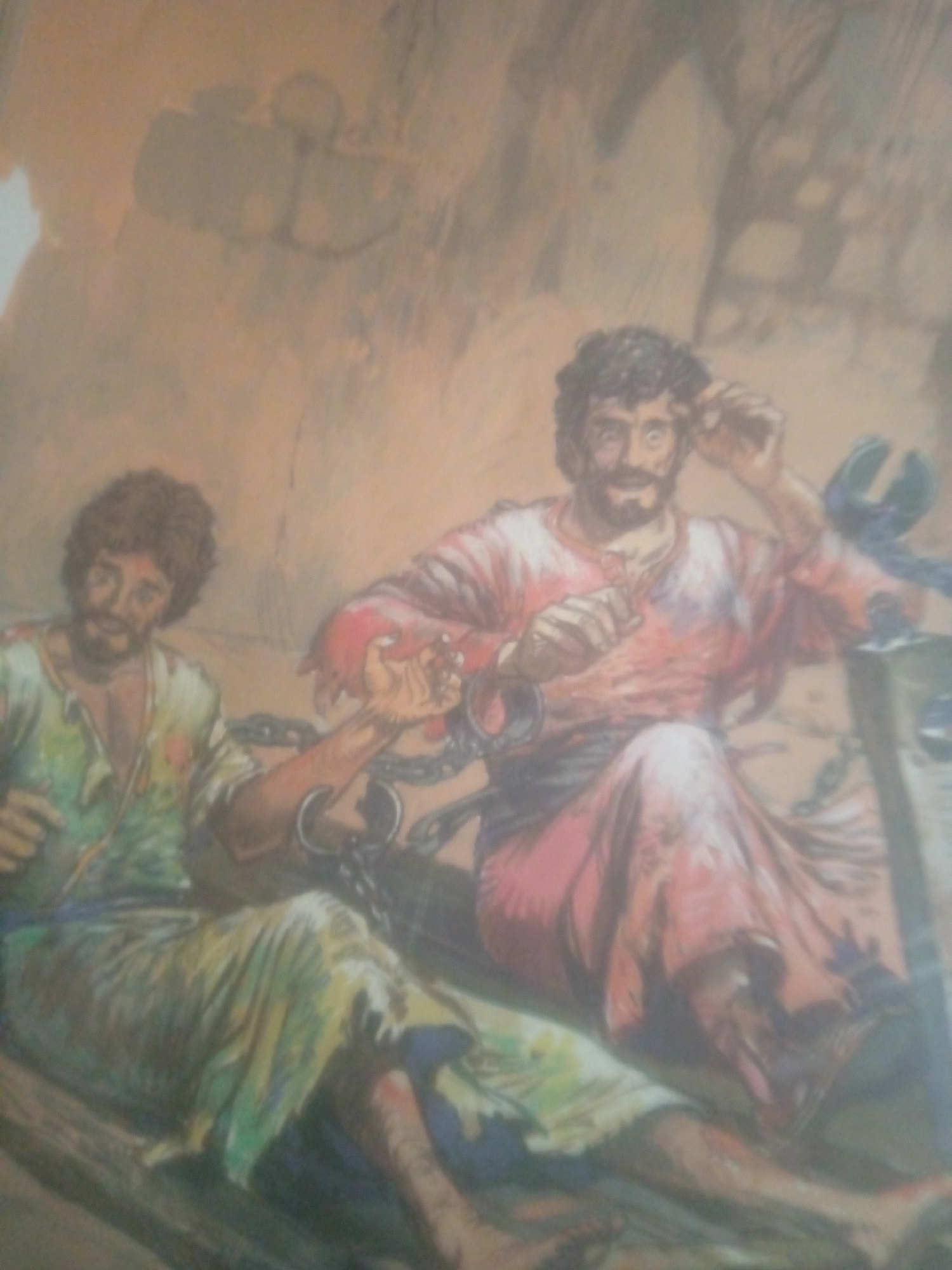 The image depicts two men in a dimly lit, possibly underground or cave-like environment. Both men appear to be bound by chains. The man on the left, wearing a green and yellow garment, is seated and looking to his left. The man on the right, dressed in a red garment, is also seated and appears to be looking up or to his right, with his hands raised. There are chains visible connecting them and possibly attached to a structure. The background is indistinct and appears to be rough, textured rock or earth. The overall impression is one of imprisonment.
