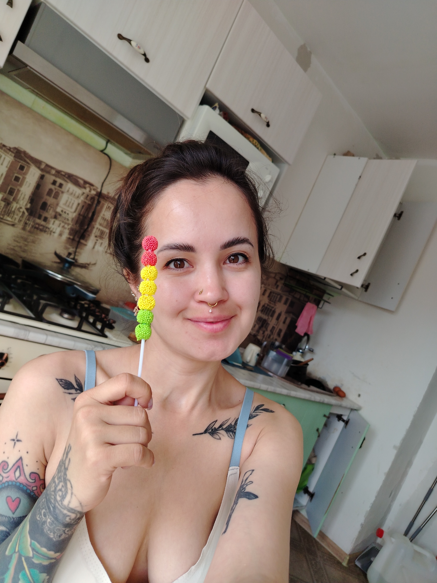 A young woman is captured in a close-up selfie within a kitchen setting in Noviy, Russia, playfully holding a colorful candy lollipop to obscure her left eye. She has dark hair, brown eyes, and a gentle smile, displaying a nose ring and a stud, as well as a lip piercing. Her chest, shoulders, and arms are adorned with numerous tattoos, including intricate floral, leaf, crown, and abstract patterns. She is wearing a light-colored bra with light blue straps.
The lollipop she holds features a white stick topped with five spherical, textured candies arranged in a color gradient: two red, two yellow, and one light green.
The background reveals a functional and lived-in kitchen. White upper cabinets with distinctive dark, decorative handles are visible, some slightly ajar. Below a range hood, a section of wall is covered with wallpaper depicting a sepia-toned scene of Venice, Italy. A white stove with black grates is partially visible. To the right, near a sink area, a pink cloth, various containers, and bottles are present. The wall on the far right is plain white, with a visible patch of repair work on the ceiling above. A white plastic jug with a red cap and cleaning tools are seen on the floor. Natural daylight illuminates the scene, and the woman's expression conveys a sense of contentment or cheerfulness. There is no visible text in the image.