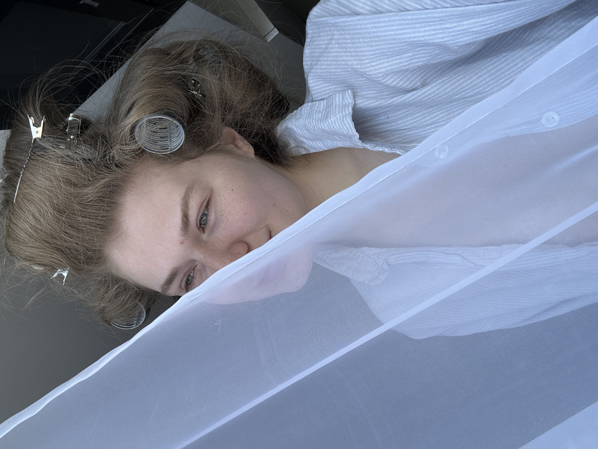 A woman with hair curlers and clips is partially visible behind a white semi-transparent fabric. She wears a light blue and white striped shirt. The scene appears to be indoors, possibly in Kazan, Russia, though the exact location within the city is not discernible from the image. A portion of a dark-colored appliance is visible in the background.