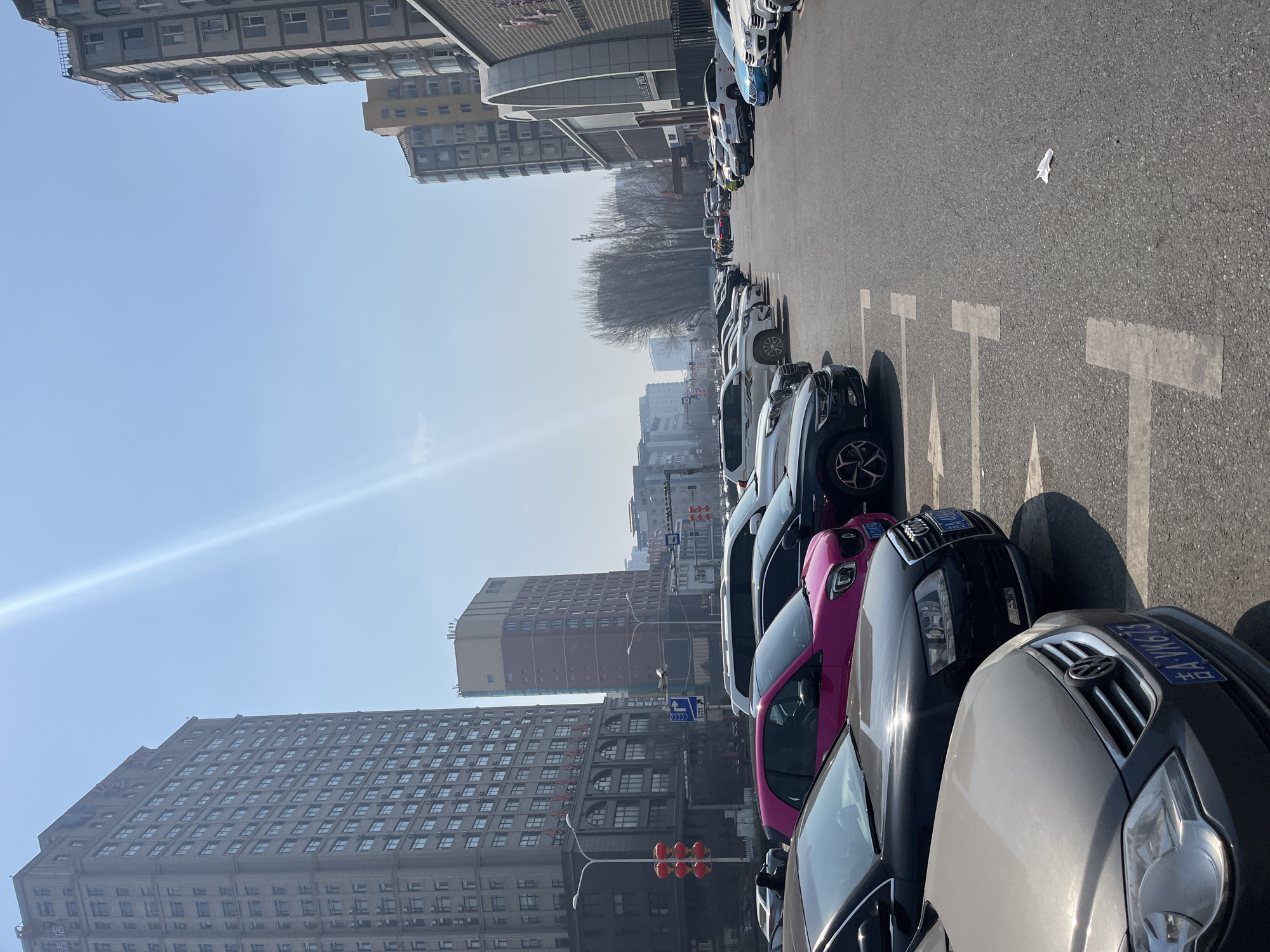 The image depicts a street scene in Changchun City, China. A row of parked cars lines the right side of the street, occupying marked parking spaces. Buildings of varying heights, including apartment blocks and commercial structures, dominate the background. The sky is blue with a slight haze. A street light with red traffic lights is visible. Based on the architectural styles, street signs, and vehicles, the location is likely within a commercial area.