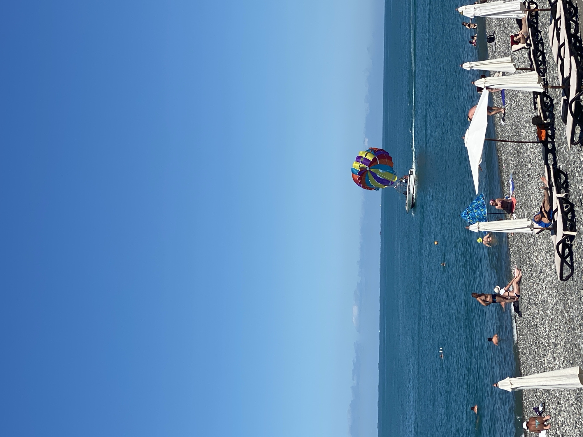 This image depicts a sunny day at a pebble beach in Sochi, Russia, likely near the Black Sea. In the distance, a motorboat tows a colorful parasail with people in it, creating a vibrant focal point against the clear blue sky and calm sea. The beach itself is populated with sunbathers lounging on chairs and towels. Several white beach umbrellas are open, providing shade. People are also seen swimming and wading in the shallow waters. The overall scene conveys a relaxed and summery atmosphere.