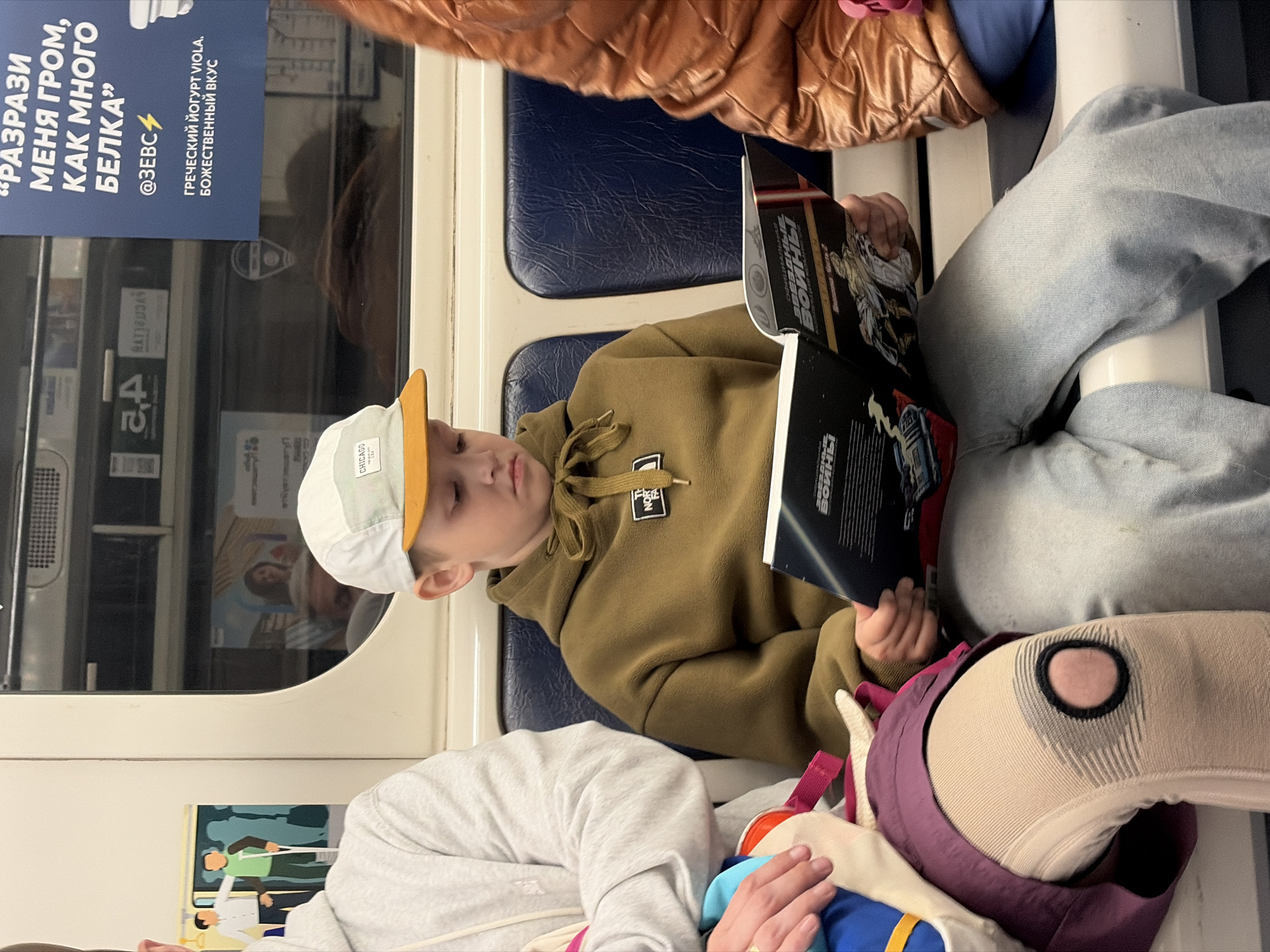 A young person is reading a book titled "Star Wars" while riding on public transportation, likely the metro, as seen by the window and seating typical of the Saint Petersburg metro system, Russia. The youth is wearing a tan hoodie with a "The North Face" logo, a white baseball cap with the word "CHICAGO" on the front, and light-colored jeans. The person is sitting next to someone wearing a brown jacket and someone holding a multi-colored bag or plush. The background contains a poster in Russian, advertising yogurt.