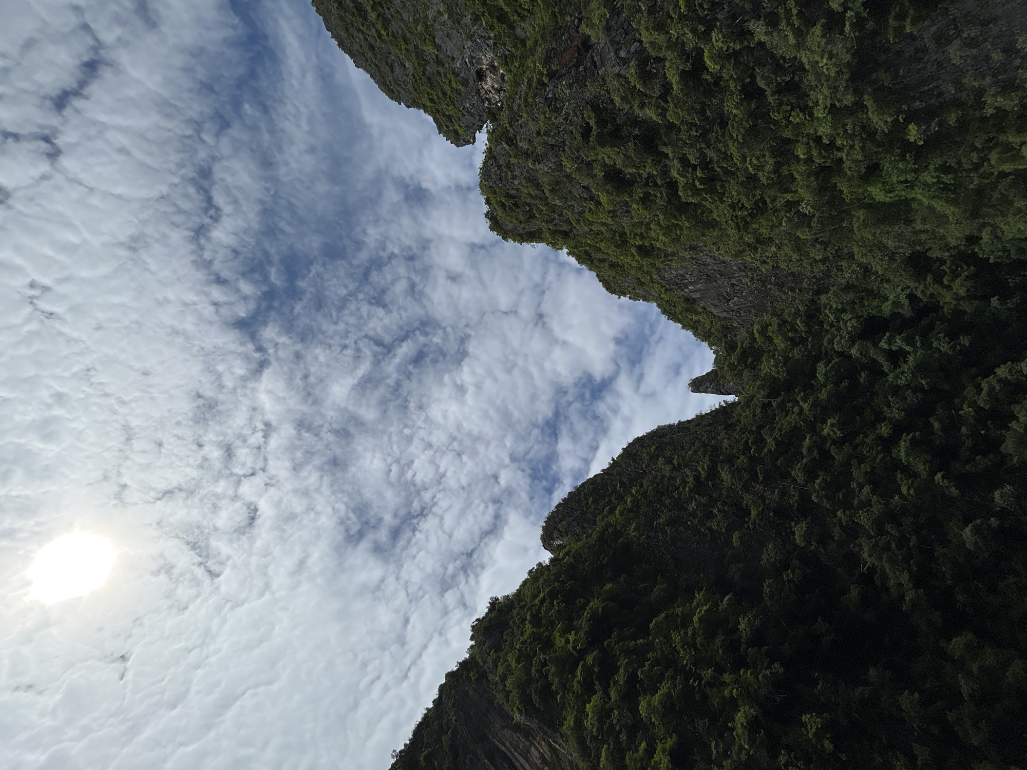 The image captures a dramatic natural landscape with two large, lush green cliffs framing a view of the sky. The sun is visible in the upper left quadrant, partially obscured by scattered, wispy clouds that create a textured pattern across the blue sky. The cliffs are densely covered in vegetation, with dark green foliage obscuring much of the rock face. The perspective suggests the viewer is positioned at a low vantage point, looking upwards between the imposing rock formations. The scene evokes a sense of natural grandeur and isolation, likely a remote or wilderness area. The location is identified as an unknown area in Thailand.