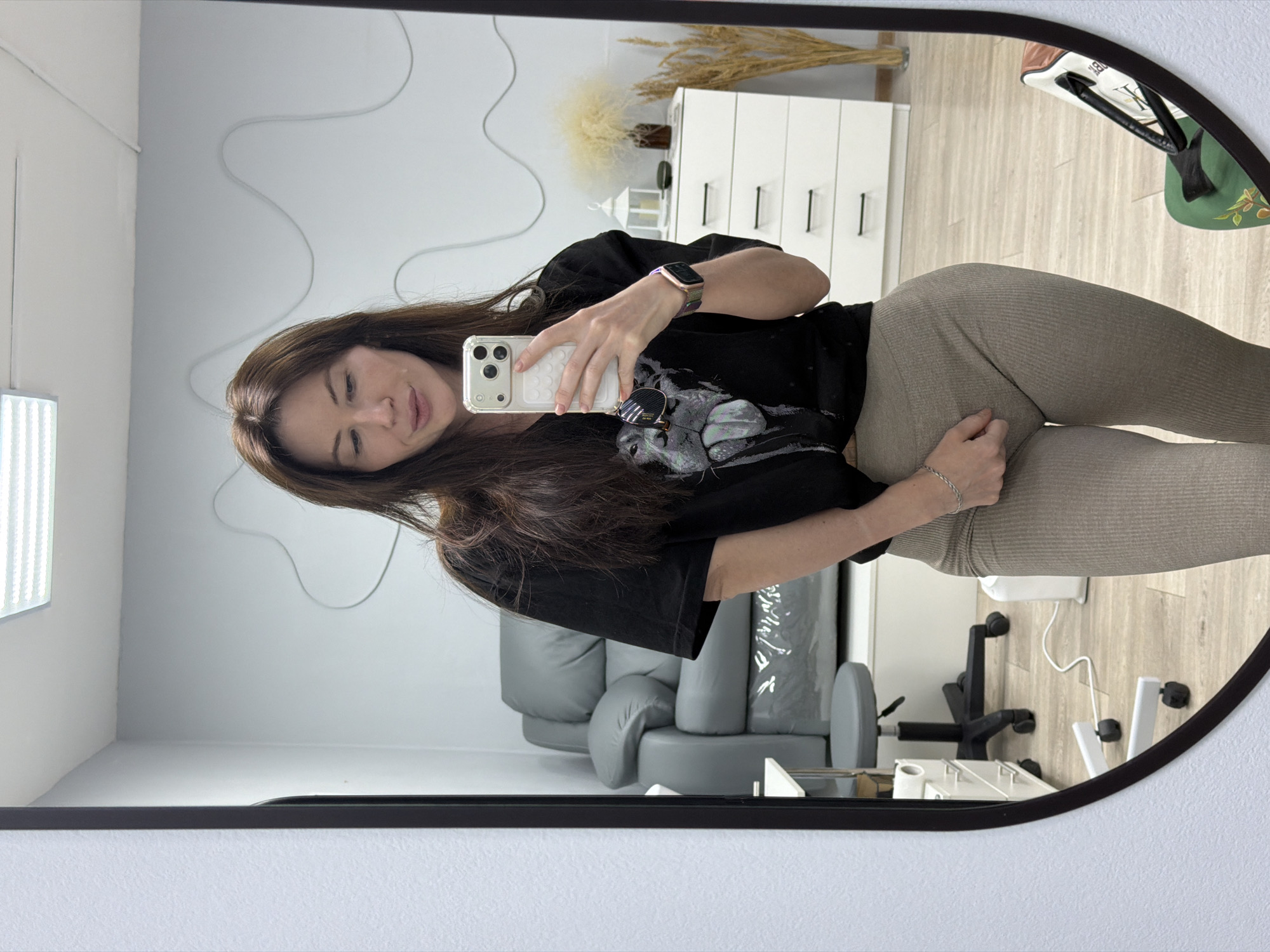 The image depicts a woman taking a selfie in front of a mirror. The woman has long, brown hair and is wearing a black t-shirt and light brown leggings. She is holding a white smartphone in her right hand. In the background, there are a white dresser, a gray sofa, and an office chair. The location could be in Perm, Russia, although there is no evidence in the image to confirm this.